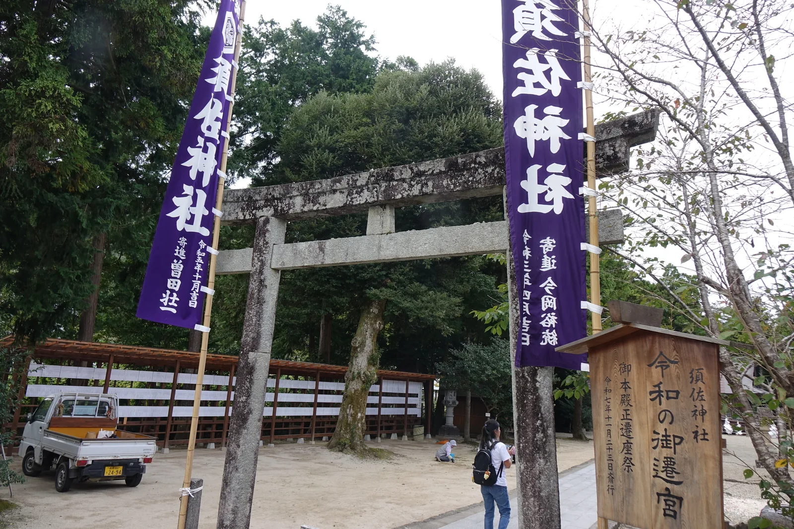 須佐神社境內入口的石造鳥居，兩側各豎一支紫色幟旗，後面是境內高大的杉，白色木柵沿著碎石地排開