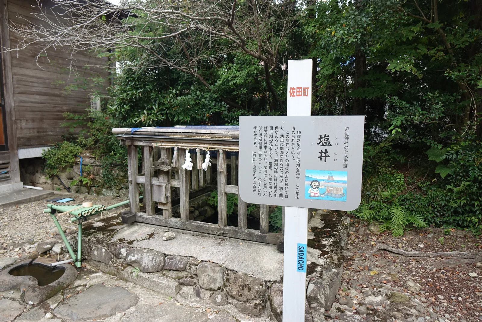 須佐神社境內的塩の井，木柵圍著，柵上垂著注連繩與紙垂，一支綠色竹管把水引到旁邊的石缽