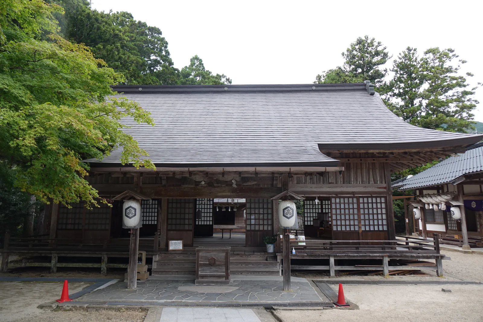 須佐神社拜殿，寬而低的切妻造，灰色屋頂往兩側緩緩張開，兩盞白紙燈籠與賽錢箱上印著六角形內含花形的神紋