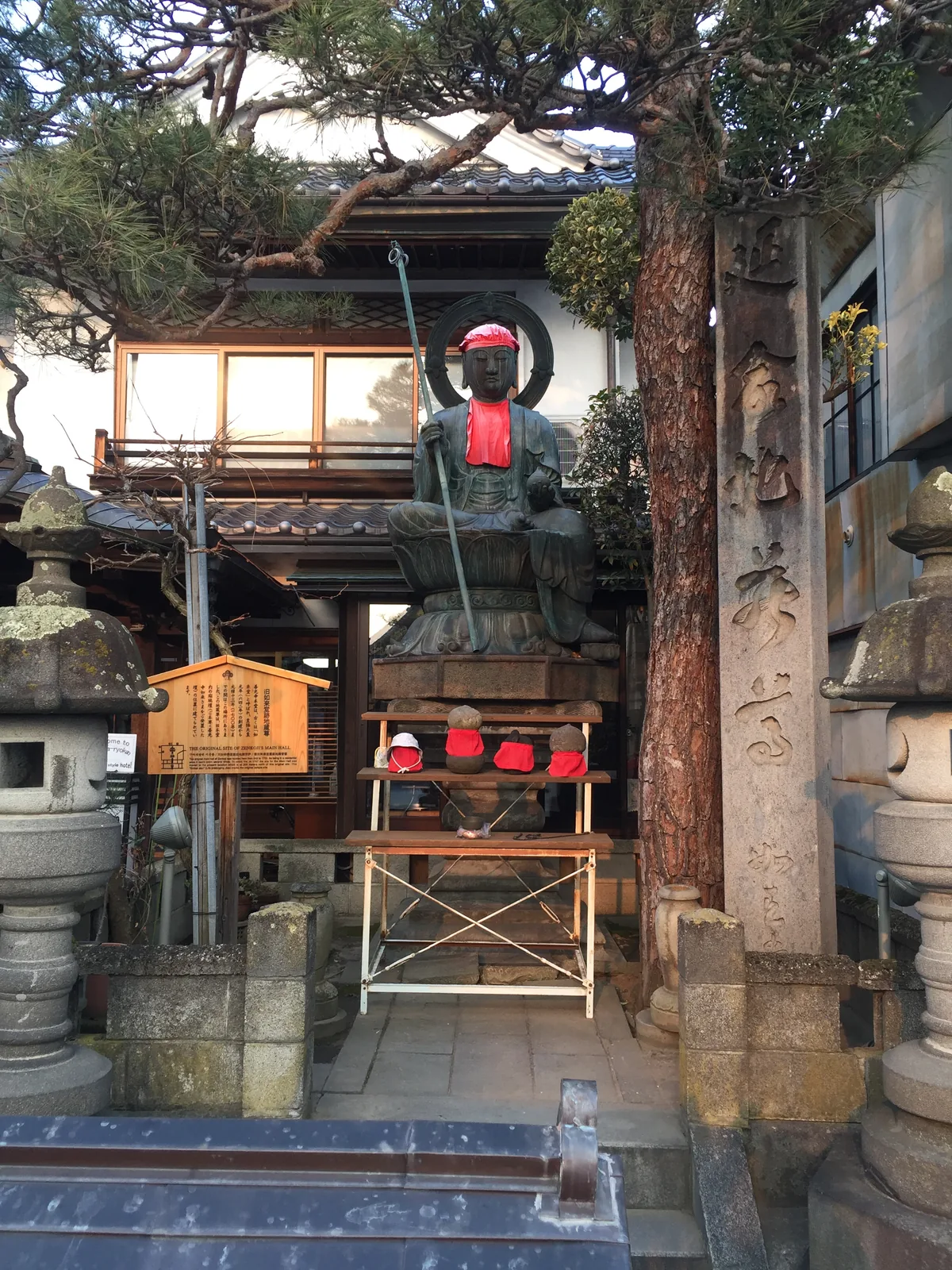 仲見世旁的旧如来堂跡地蔵尊：戴紅帽、掛紅圍兜的延命地藏坐在石臺上，左側立著說明舊本堂位置的木札，右側是刻字的石柱與石燈籠