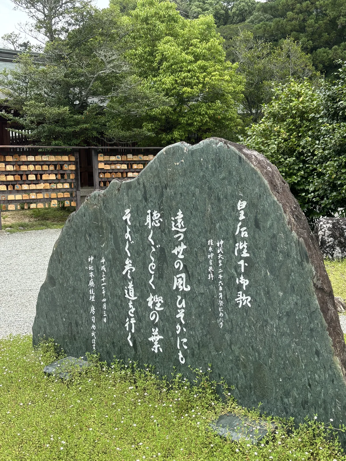 同一首歌刻在一塊深綠色的自然石上，草書縱書。旁邊是繪馬掛所，掛滿的木牌在背景裡連成一片。
