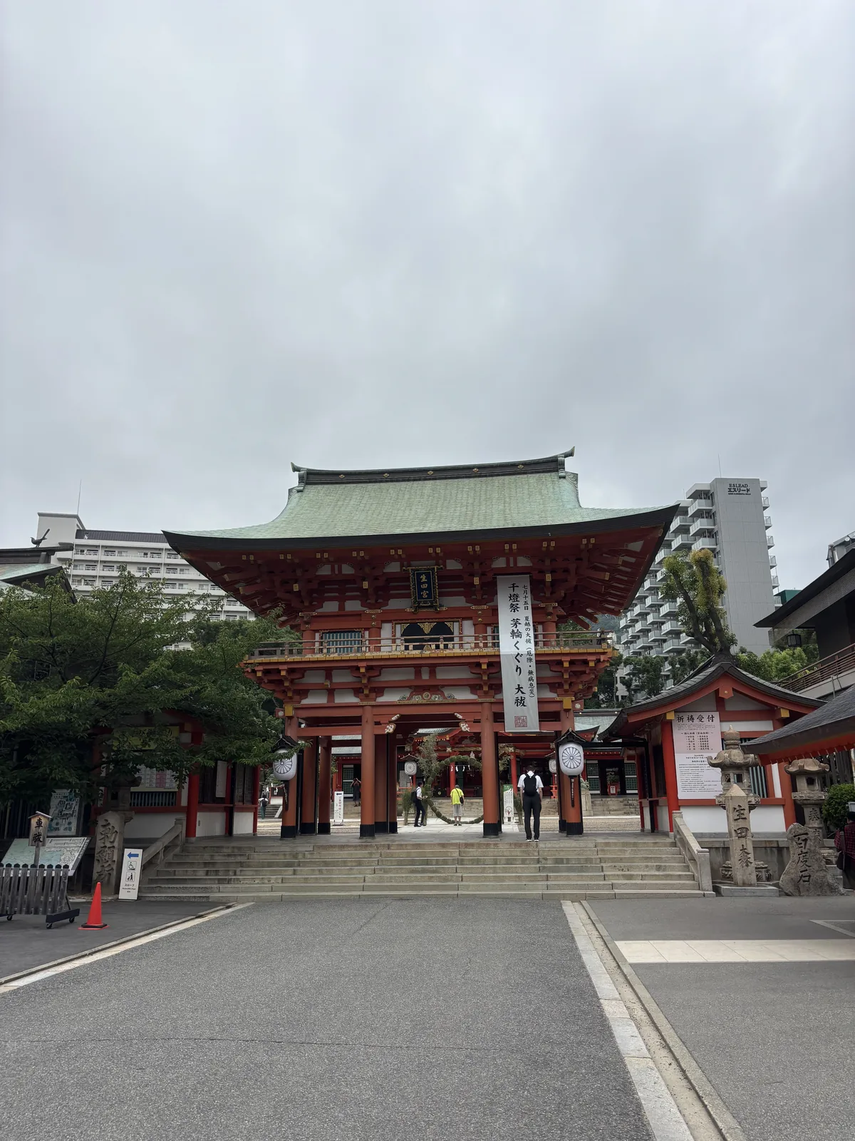 樓門正面，隔著參道與石階看。朱塗的柱與斗栱，屋頂是銅板葺、泛著綠青，入母屋造的曲線；門前左右立白色的幟旗。背後探出神戶的公寓大樓——這座神社在三宮的街正中央。