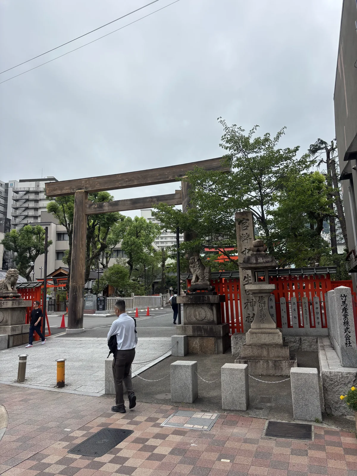 素木大鳥居橫在下山手通旁的人行道盡頭，右側是刻「生田神社」的社號標，基座刻著奉獻的株式會社名。地面是磁磚鋪的人行道，柱腳邊就是車道與交通錐。參道的第一步在馬路邊上。