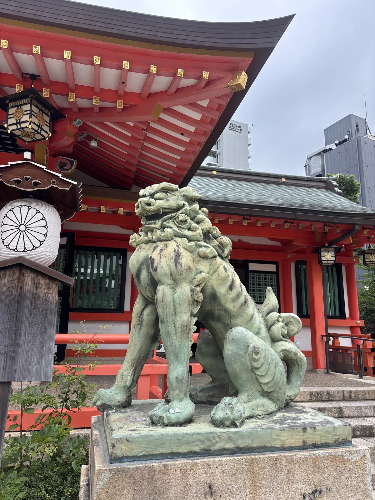 一對狛犬的其中一隻，坐在花崗岩台上，通體綠鏽。背後是樓門的朱色與綠青屋頂，簷下吊著銅燈籠，一盞畫菊紋的白燈籠掛在旁邊。