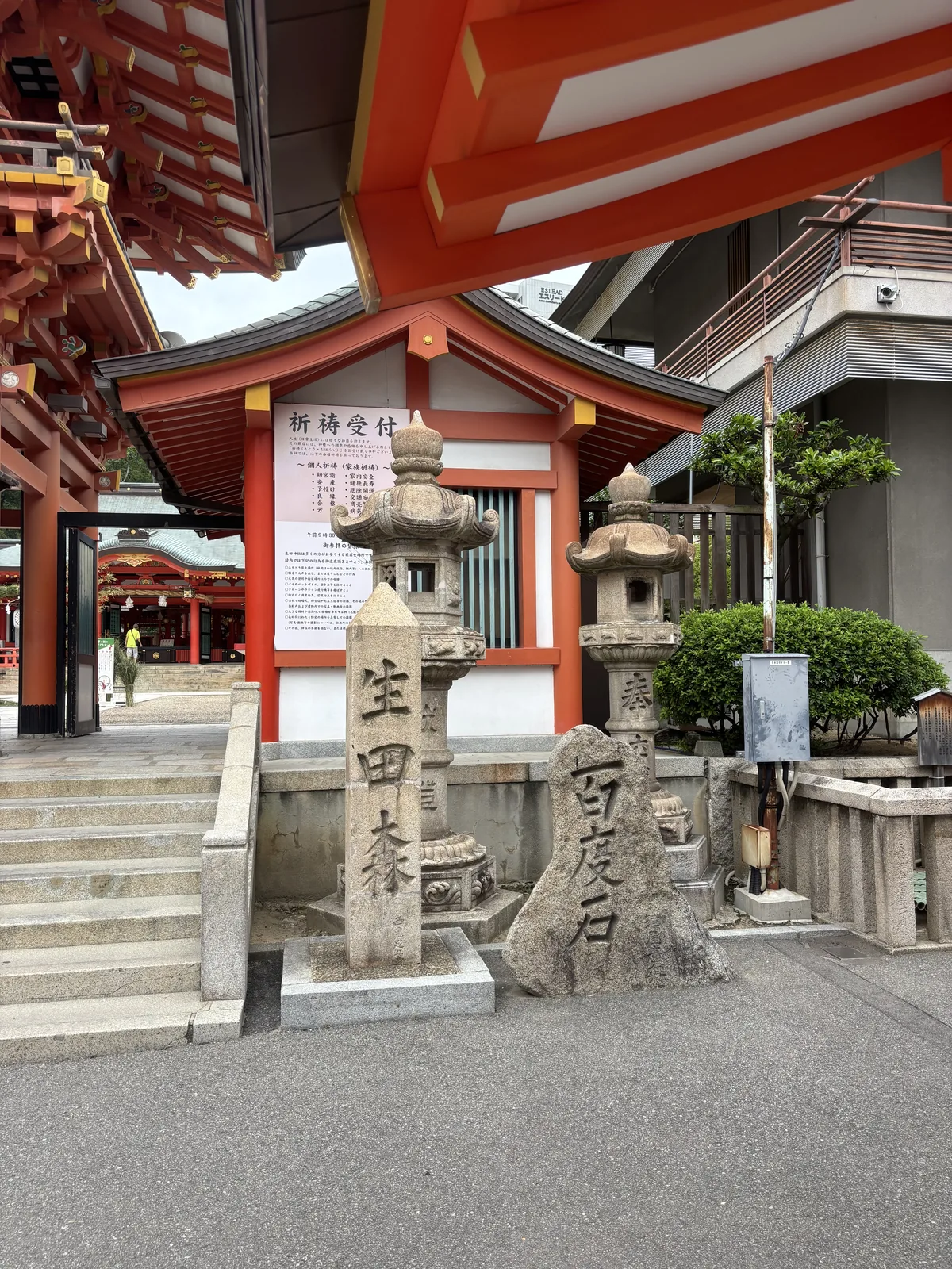 兩根石柱並立：右刻「生田森」，左邊那塊自然石刻「百度石」——百度參拜時往返計數用的標石。後方是朱塗的祈禱受付，掲示列著初宮詣、安產、子授け、良緣、家內安全、商賣等項目，受付從上午九點半開始。