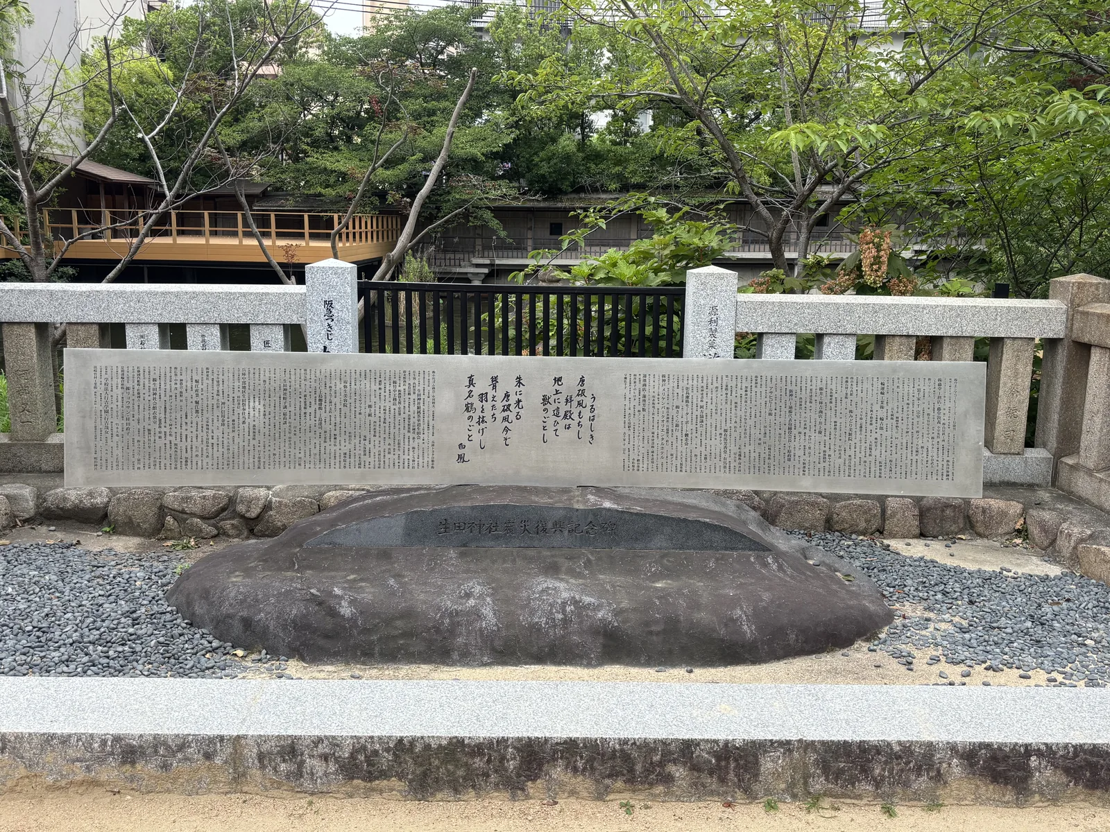 一塊橫臥的自然石，正面嵌黑石刻「生田神社震災復興記念碑」；後方的長條石板刻著捐納者的名字與一首和歌。碑立在鋪了黑色礫石的方框裡，背後是石造欄杆與樹。