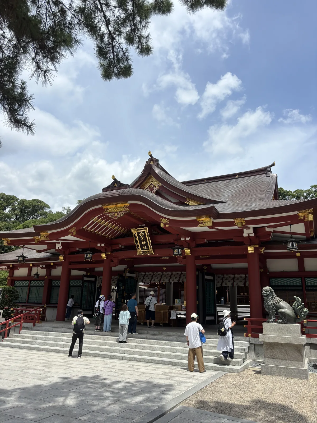 拜殿正面。朱塗、深褐色的屋面，中央一具大唐破風的向拜，金色的懸魚與飾金物排在朱色上；正中高掛金地扁額「西宮神社」——大榮創業者中內㓛在日本零售業首次達成年營業額一兆日圓的紀念參拜時奉納的。右側石台上一隻狛犬，簷下吊著燈籠。