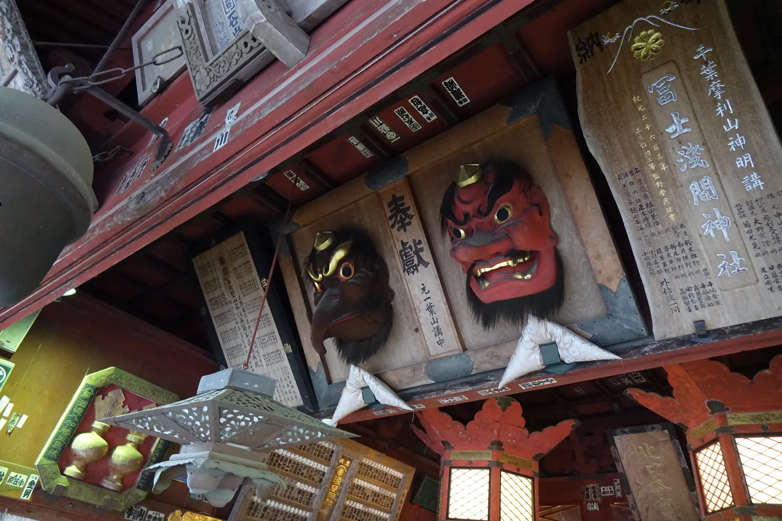 天井底下的橫木上釘著兩張面具：右邊朱紅、金牙、怒目的大天狗，左邊深褐、長喙、有鬍的烏天狗。兩者之間的木板寫「奉獻」，落款「元一葉山講中」。右側一整片欅木刻「千葉摩利山神明講／冨士淺間神社」，下方密排奉納者名。