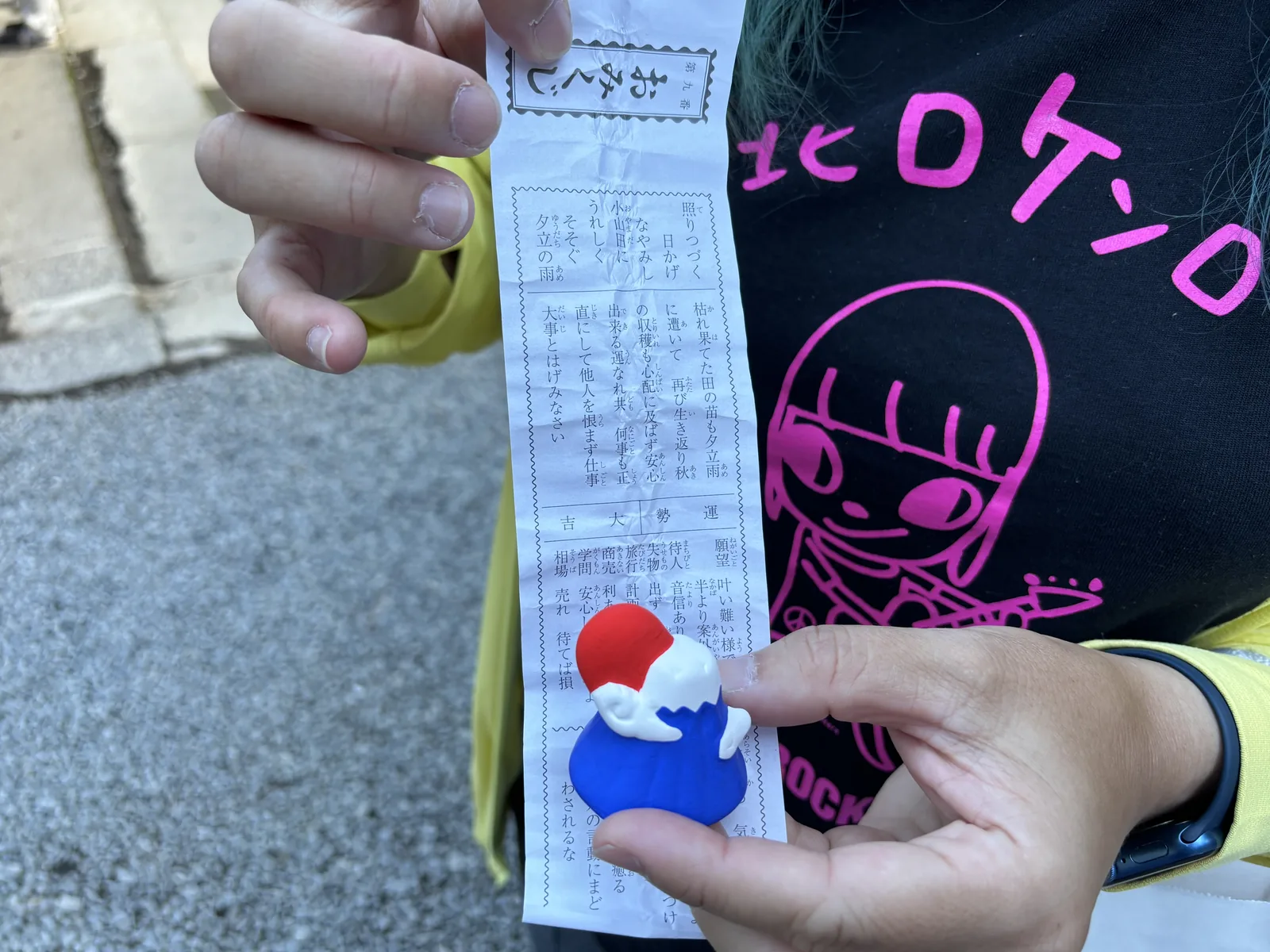 手上攤開一張御神籤，旁邊放著一枚做成富士山形狀的小土鈴：藍色的山體、白色的雪冠、紅色的頂。這是這座神社把「御神體是那座山」講給參拜者的最短一句話。