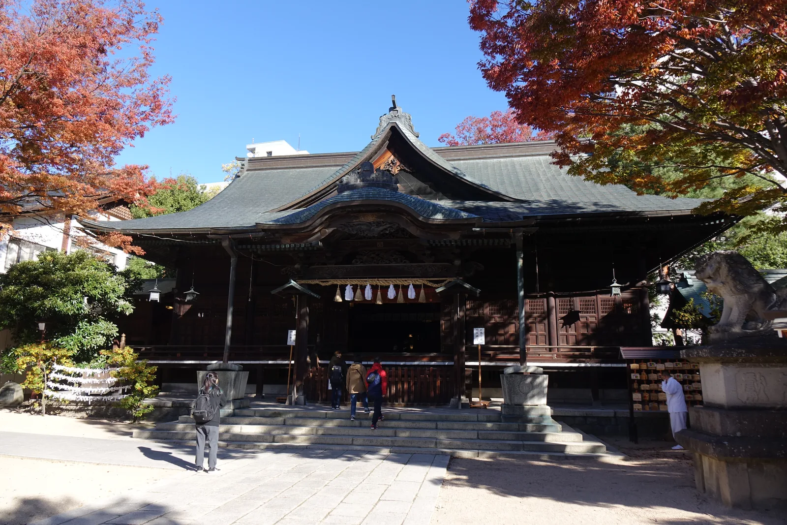 拜殿的正面。入母屋造（いりもやづくり，即歇山頂），木料風化成深褐，正面完全開放，簷下橫過一道粗的注連繩並垂下白色紙垂。左側可以看到石造的狛犬與其台座。屋頂兩坡之間的紅葉正紅。這一棟是大正十三年（1924）重建的。