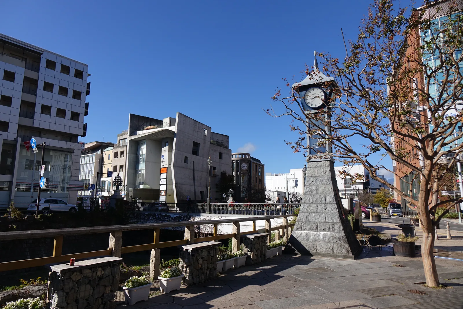 從橋上往東看。河道被石砌的護岸夾住，河床窄而淺，兩岸是市街的背面與現代的大樓。右岸立著一座石造的時計台。這條河把松本的城下町切成南北兩半。