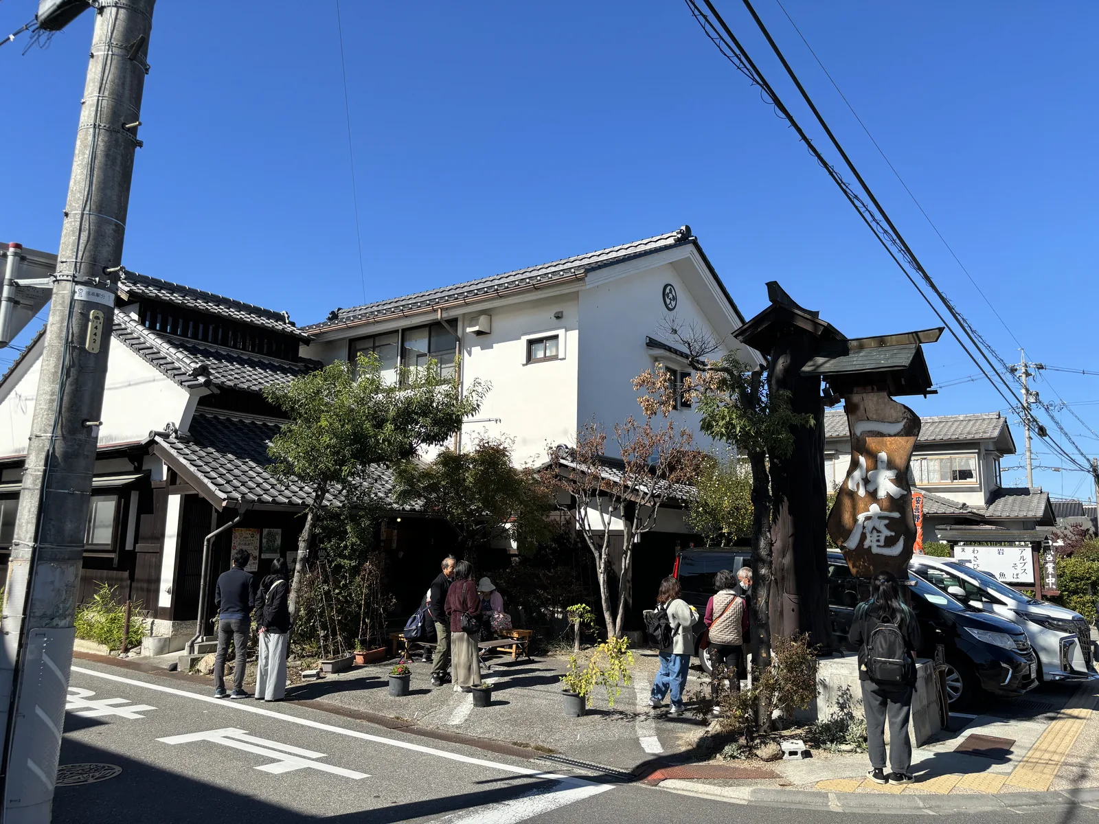 白牆黑瓦、外牆塗著厚灰泥的町家沿街排開，路口立著一支黑色的木招牌柱，柱上刻著店名。店門前站著一排等位的人。這條就是驛前通り，神社的鳥居在它的一側，往東走就是田。