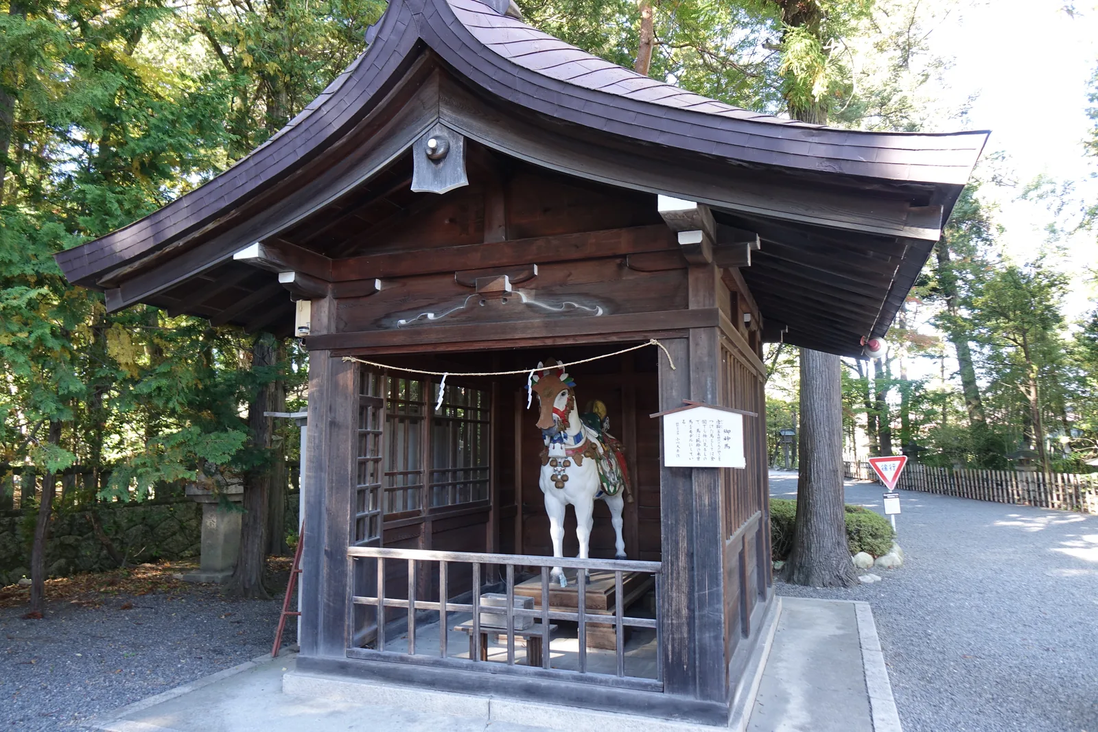 一座獨立的深色木造小屋，屋根的曲面平緩，正面與側面設木柵，簷下橫著一道注連繩。裡面立著一匹白色的神馬像，配著彩色的鞍與轡。右柱上掛著一塊白紙的說明。