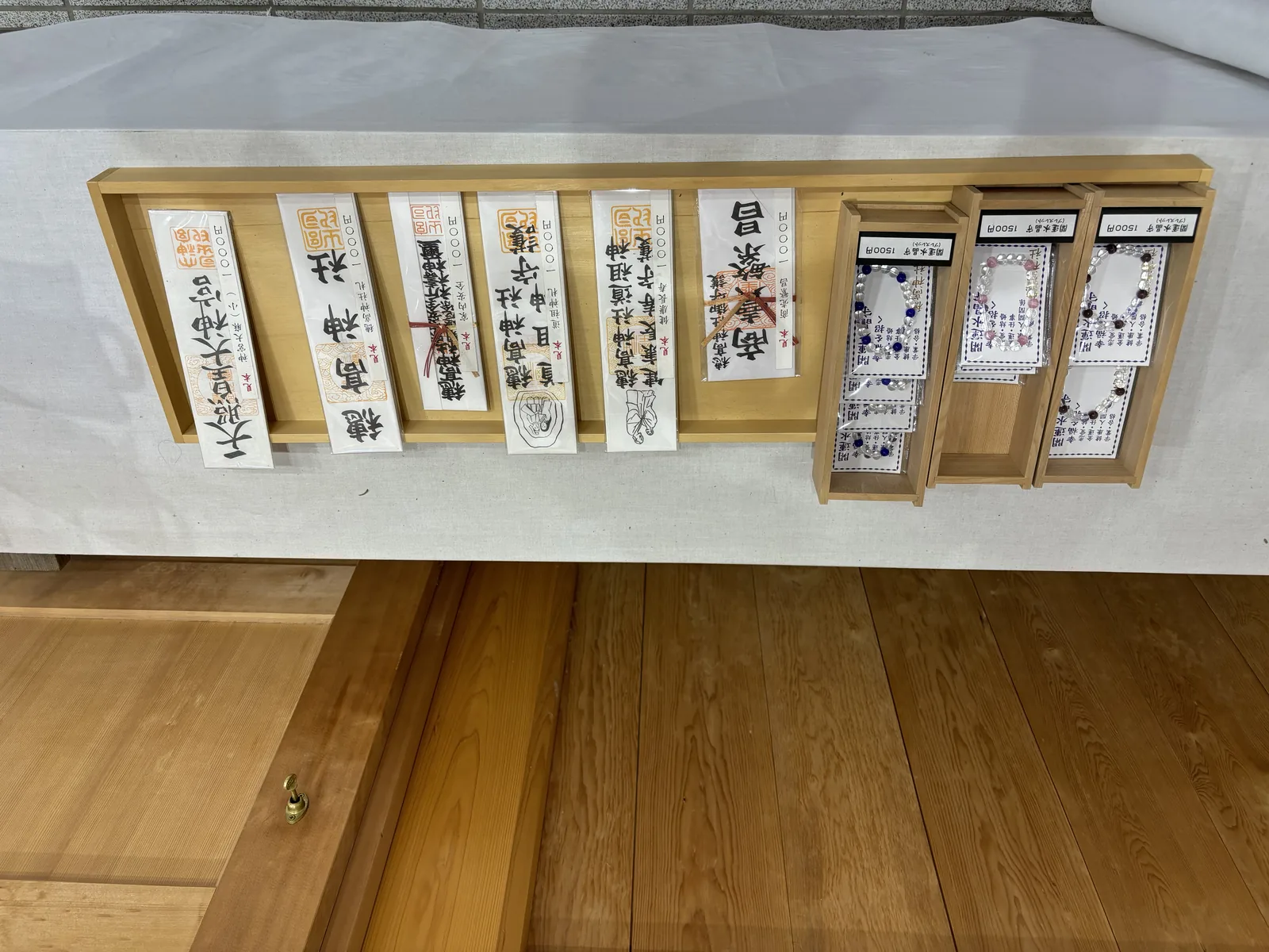 白布鋪的長桌上，木盤裡並排著一列神札：右起「天照皇大神宮」「穗高神社」，往左依次是「穗高神社神璽」「一目神守護」「建築起工守護」「穗高神社道祖神」與「商賣繁昌」。左半是幾盒裝袋的御守。木札全是素木墨書。