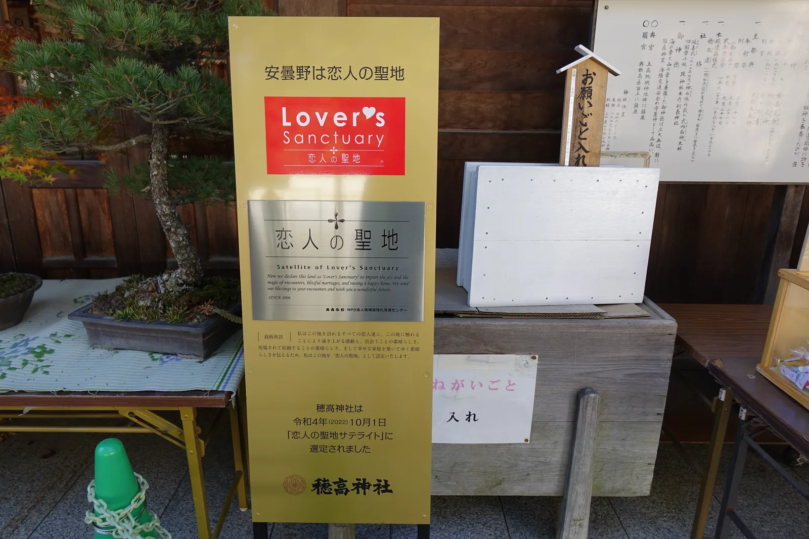 立在社務所前的一塊黃底看板。上端橫寫「安曇野は恋人の聖地」，意思是安曇野是戀人的聖地；中央是紅底白字的「Lover's Sanctuary 恋人の聖地」標誌，下方的說明文末尾寫著：穗高神社於令和四年（2022）十月一日被選定為「恋人の聖地サテライト」。旁邊擺著盆栽，以及一個寫著「お願いごと入れ」（O-negaigoto-ire，投願望的箱子）的投函箱。