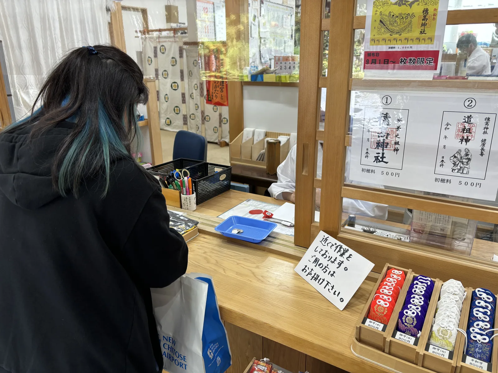 社務所的御朱印窗口。玻璃上貼著兩張御朱印的樣張，①穗高神社、②道祖神，下方各標初穗料五百圓；上方是一張黃底、畫著船與浪的御船祭限定御朱印，紅條寫「9月1日～ 枚数限定」。窗口前一位參拜者正在填寫，台面右側排著紅、紫、白、藍四色的御守。
