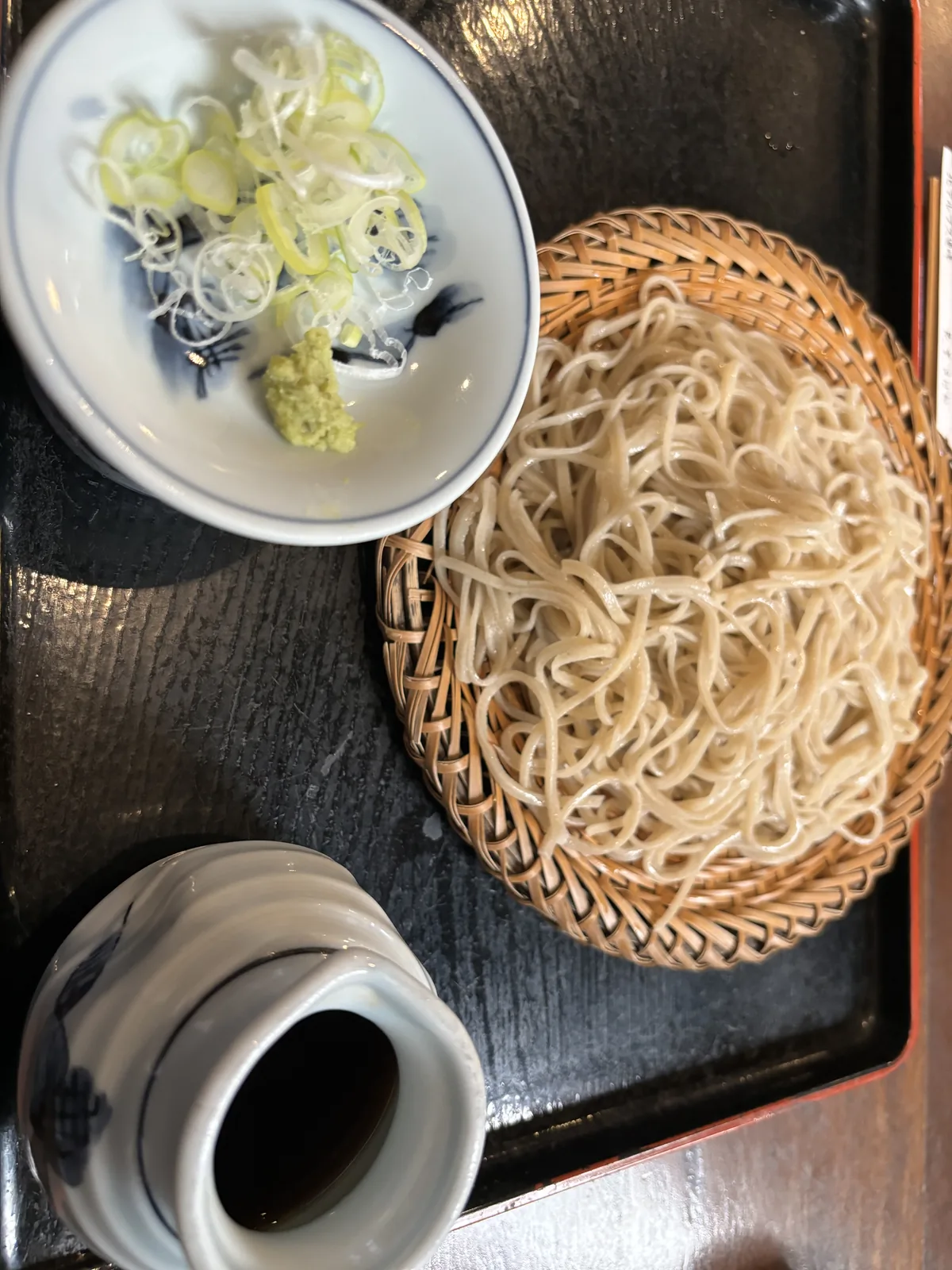 竹笊上一束細切的蕎麥麵，旁邊一小碟蔥花與山葵，前方一只盛著沾汁的染付瓷壺，全放在朱漆的托盤上。