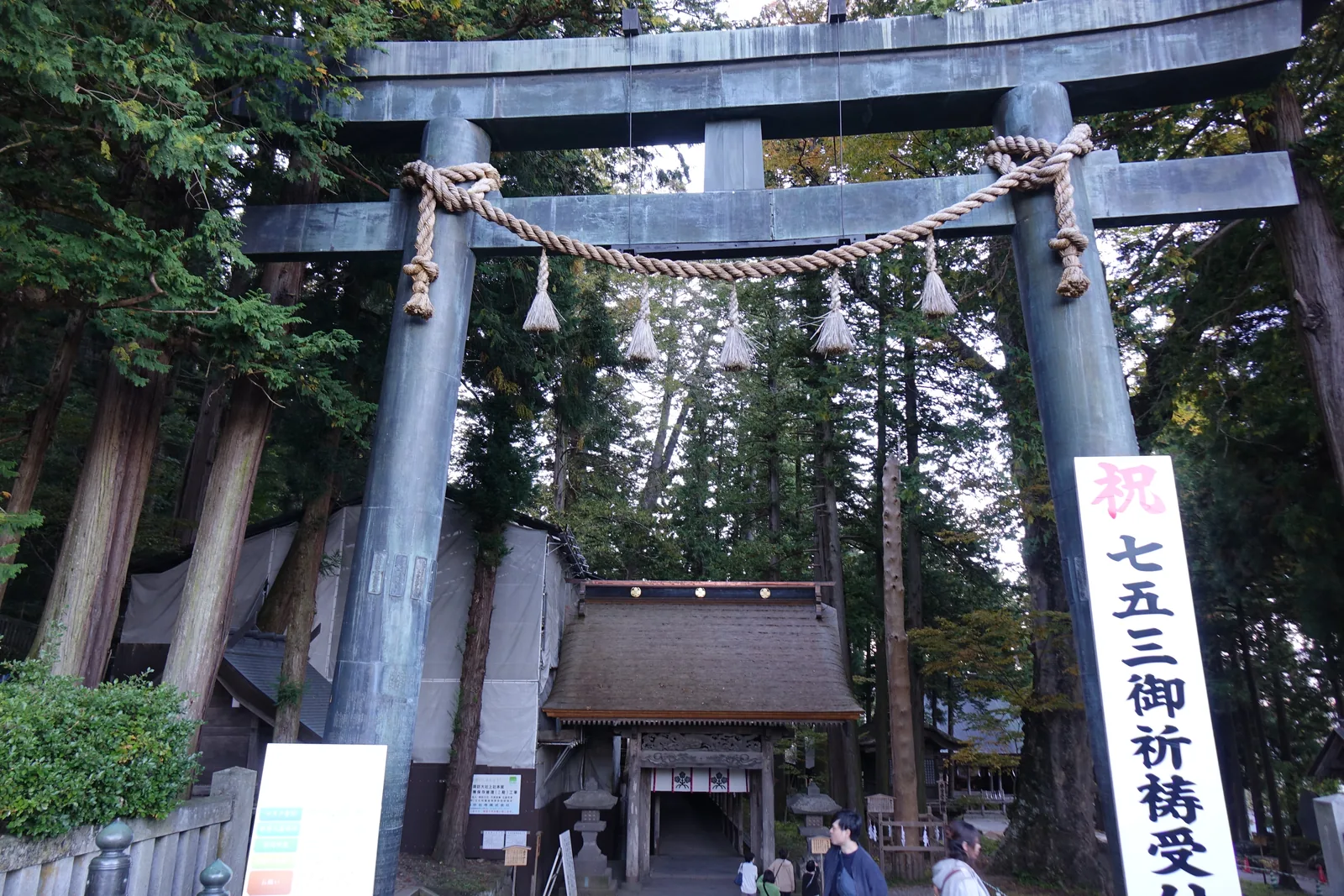 石造的大鳥居橫過畫面，笠木上掛著一道注連繩（しめなわ shimenawa，標示神域的稻草繩）。鳥居的右柱旁立著紅底白字的「祝 七五三御祈祷受付」幟，穿過鳥居可以看見境內的建築與參拜的人。