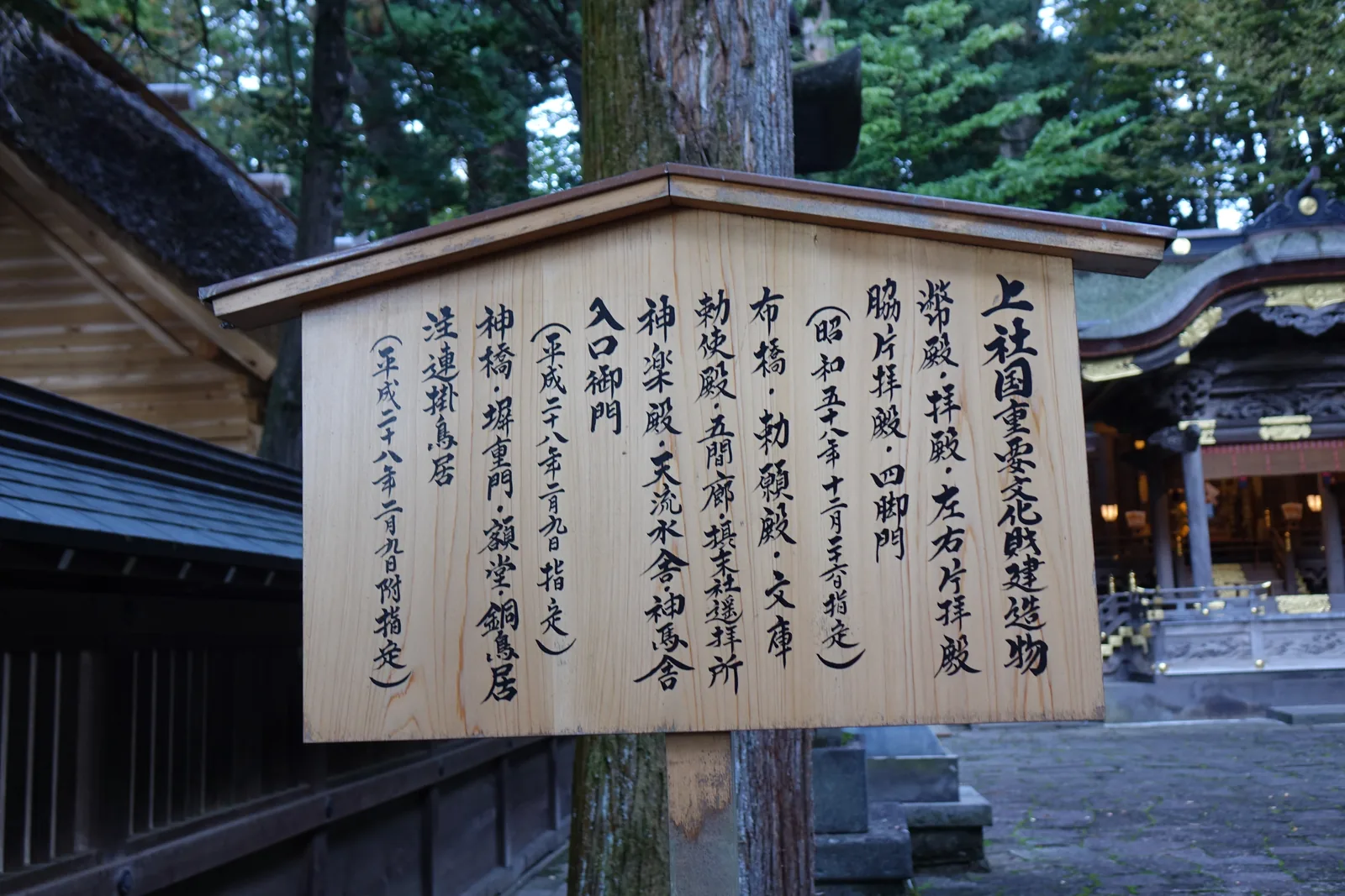 一塊帶小屋頂的杉木立牌，墨書「上社国重要文化財建造物」，底下分三批列出：幣殿・拝殿・左右片拝殿・脇片拝殿・四脚門（昭和五十八年指定）；布橋・勅願殿・文庫・勅使殿・五間廊・摂末社遙拝所・神楽殿・天流水舎・神馬舎・入口御門（平成二十八年指定）；神橋・塀重門・額堂・銅鳥居・注連掛鳥居（同日附指定）。右後方就是幣拜殿的簷角。