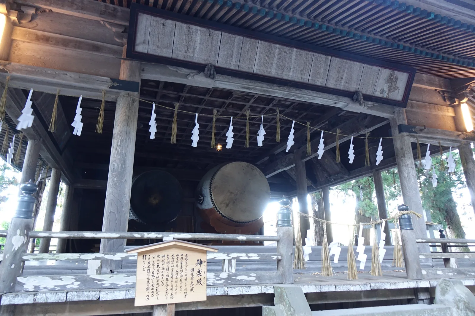 從側面往神樂殿內看。簷下垂著一整排紙垂（しで shide，摺成階梯狀的白紙），殿內的深處靠著一面巨大的太鼓，鼓面朝向鏡頭；前方掛著一塊小小的木製說明牌。這面江戶時代做的太鼓直徑一公尺八十。