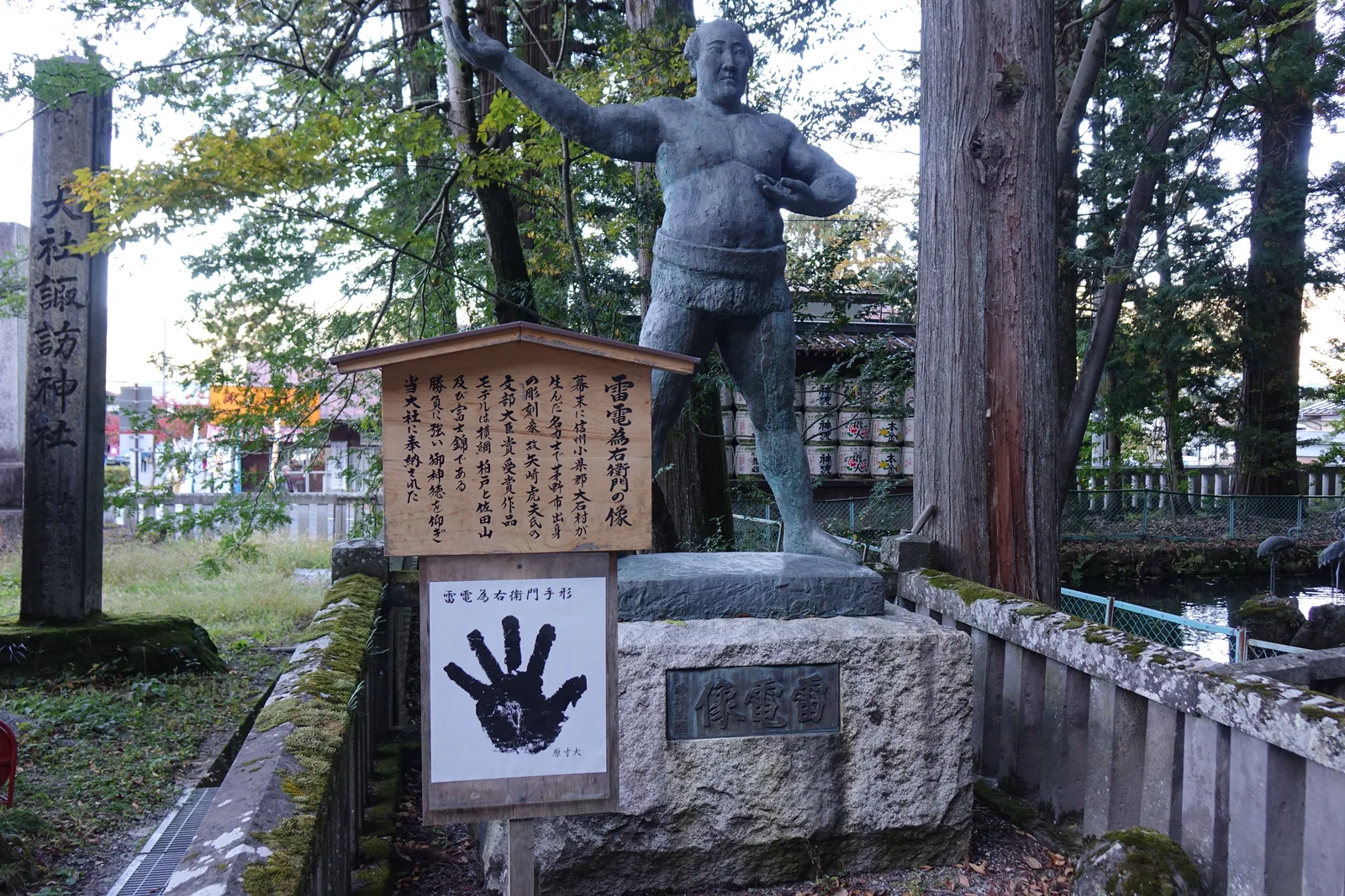 石臺上一尊力士的銅像，右手高舉，臺座刻「雷電像」。旁邊立著說明牌，牌下另掛一塊白板，印著「雷電為右衛門手形」——一個墨黑的手掌拓印。最左邊是社號標的石柱。