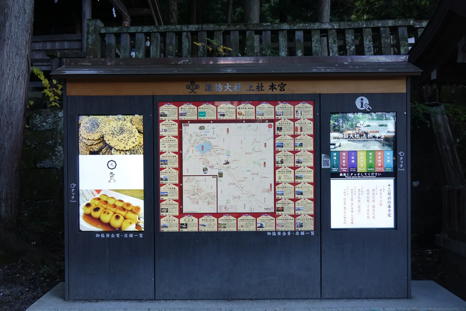 一座黑色的戶外看板，頂上木牌題「諏訪大社 上社本宮」。中央是門前一帶的商家地圖，兩側各一片可觸控的螢幕，右側那片列著當月的行事預定，左側那片輪播著門前店家的甜點照片。