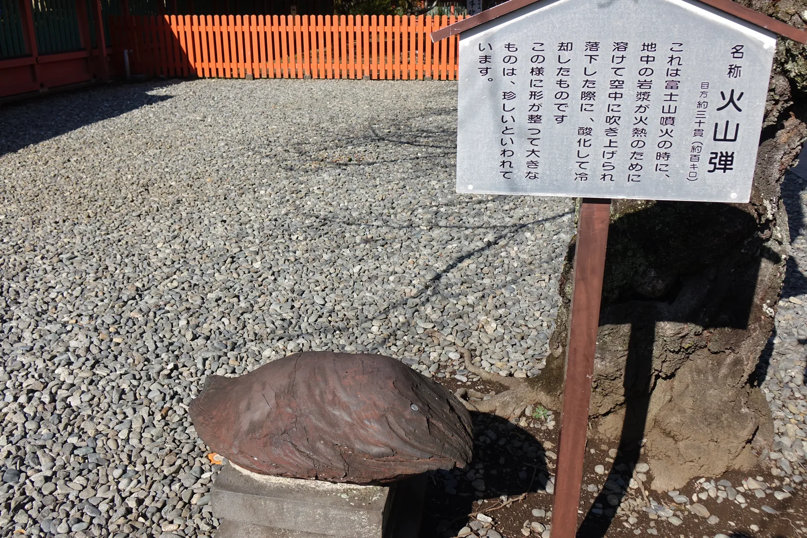 火山彈與立牌的近景。牌上直書「名称　火山弾」，右下小字「目方約三十貫（約百キロ）」——重約三十貫，牌上自己換算成大約一百公斤；內文說明它是噴火時地中的岩漿被吹上空中、落下時氧化冷卻而成，形狀這麼整齊又這麼大的很少見。石頭本身像一枚壓扁的水滴。