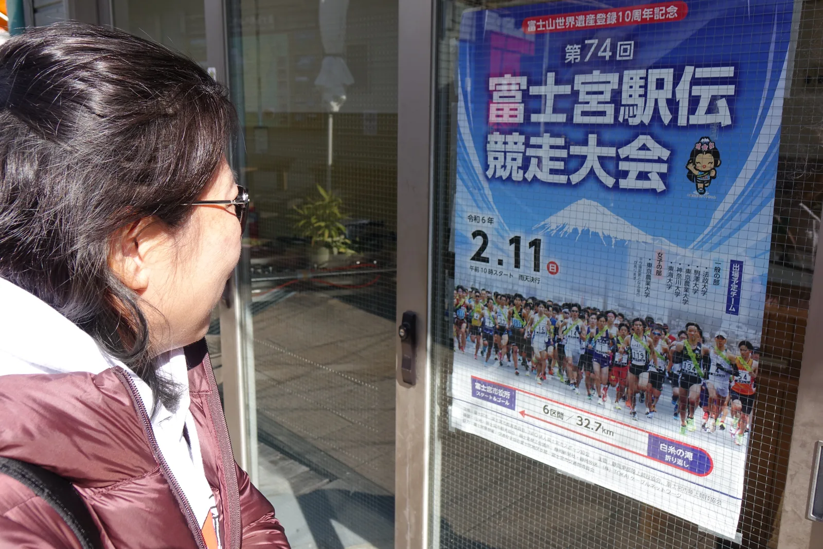 玻璃門上貼著一張藍底的海報，主標「第 74 回　富士宮駅伝競走大会」，頂端一行「富士山世界遺産登録 10 周年記念」，下方寫日期「令和 6 年 2.11（日）」與「6 区間／32.7km」，起終點是富士宮市役所、折返點是白糸の滝。一位參觀者側著臉在看。