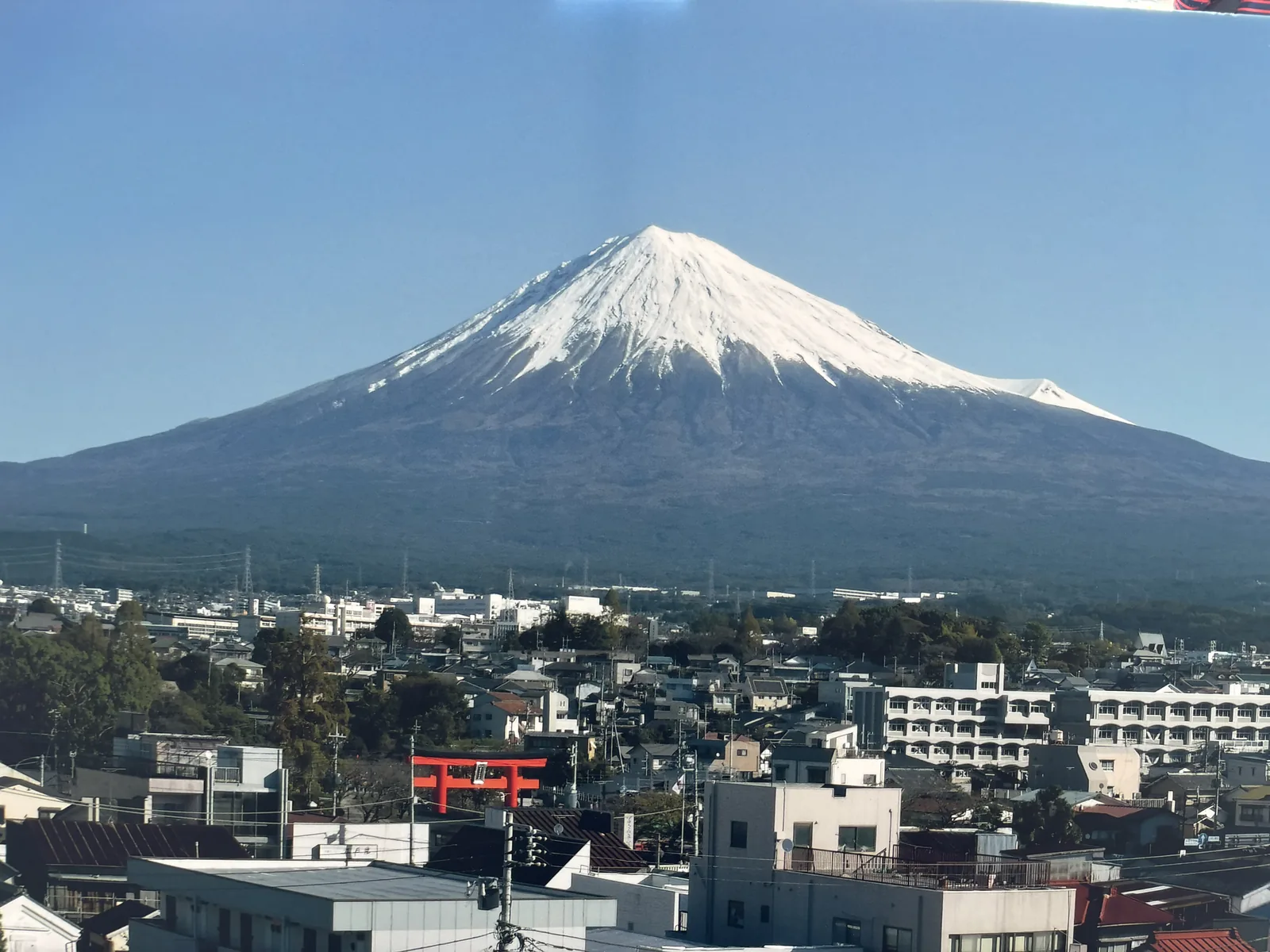 從中心上層的展望室往北看。前景是富士宮的市街屋頂、電線與一座朱色的大鳥居，後方富士山完整地出現，山頂積雪，山體幾乎沒有雲。同一天中午在櫻の馬場上，這座山被積雲整個遮住。