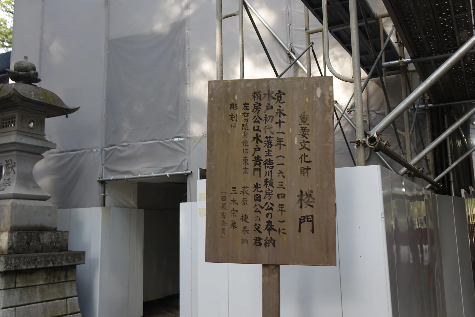整座門被鷹架與白色帆布包住，只剩底下一條通道。前面立著木牌：「重要文化財　樓門／寬永十一年（一六三四年）に水戶初代藩主德川賴房公の奉納」——寬永十一年由水戶初代藩主德川賴房奉納；牌上並記賴房是水戶黃門光圀的父親，左右的隨身像由東京的萩原捷奉納，雕刻出自三木宗策。這是「令和的大改修」四棟裡的最後一棟，二〇二四年動工。