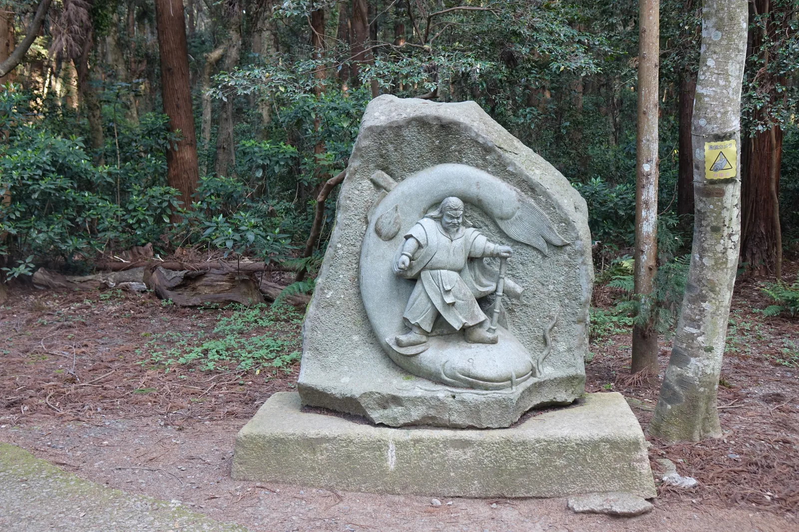 一塊自然石上刻著武甕槌大神踩住大鯰的圖：神拄著一根棍，一腳踏在鯰背上，鯰的身體繞成一圈把人框在中間。地底深處的要石壓著引起地震的鯰——這個說法在江戶時代流傳極廣，這塊碑把它做成了看得見的東西。