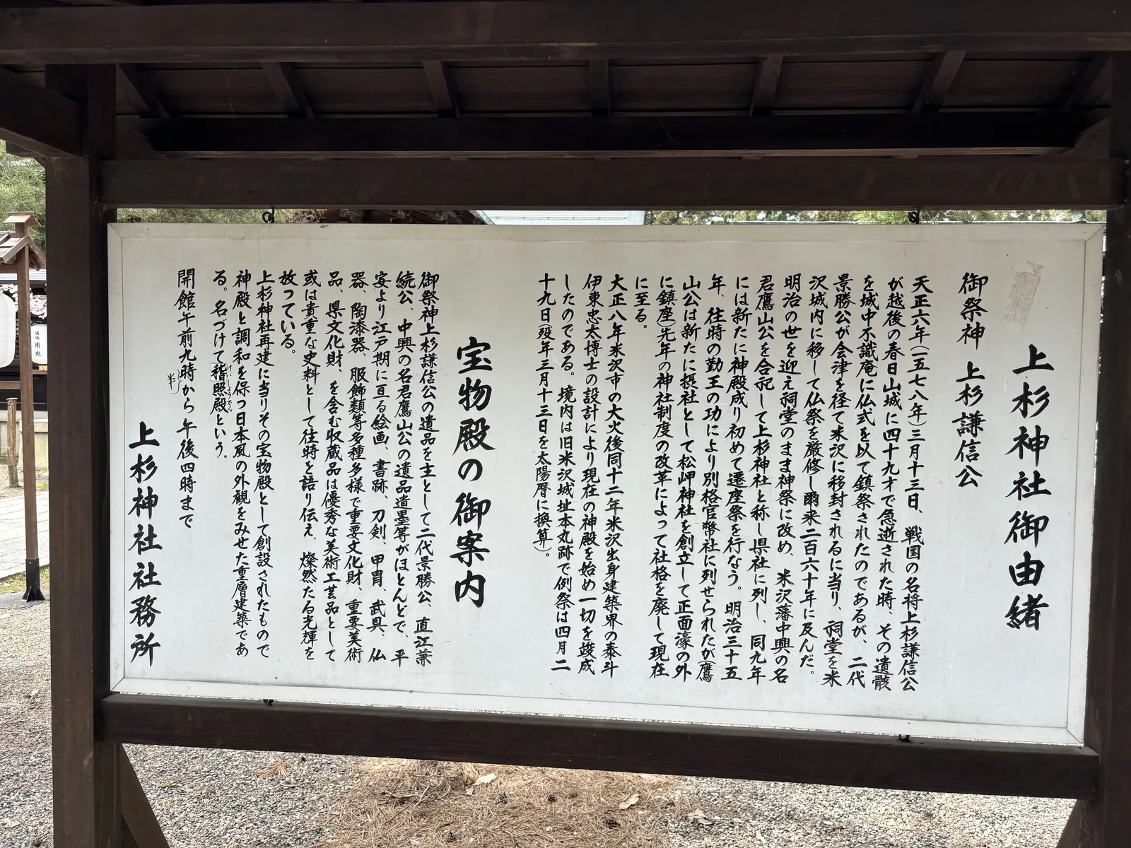屋根付的木框裡一面白色說明板，直排毛筆體，標題「上杉神社御由緒」，右起第一行寫「御祭神　上杉謙信公」，左半另闢一欄「宝物殿の御案内」，落款「上杉神社社務所」。祭神欄只有一個名字——一九〇二年之後就是這樣了。