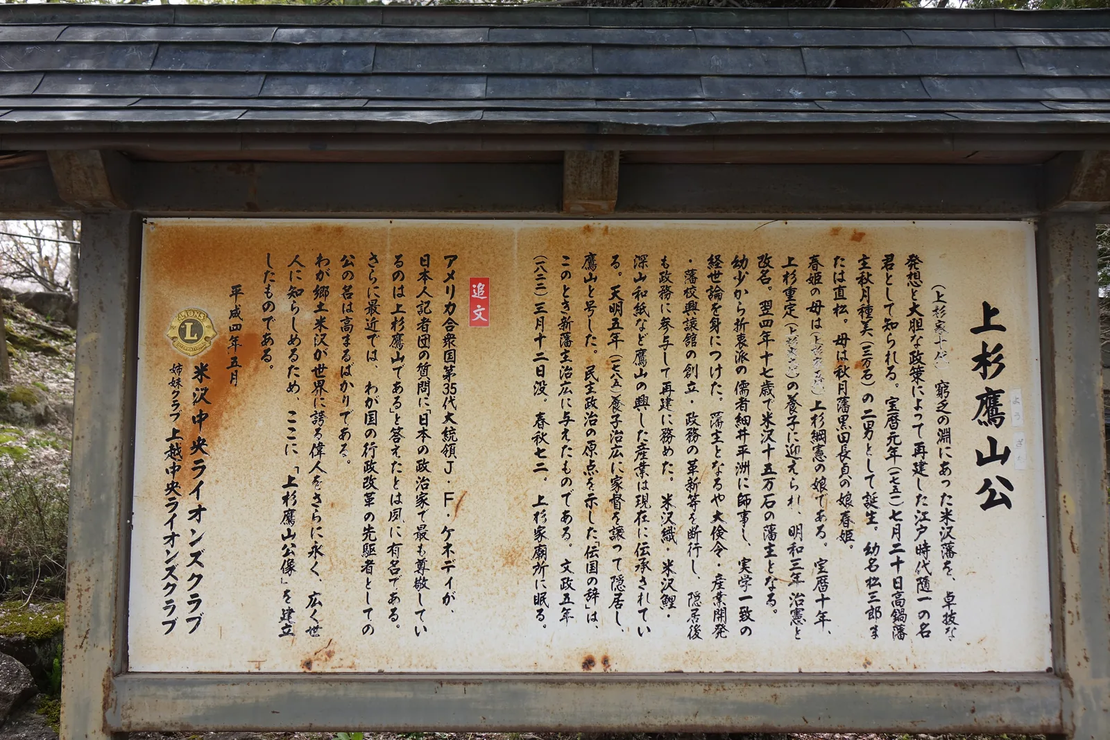 瓦屋根下一面鏽跡斑斑的白色說明板，直排文字，右起標題「上杉鷹山公」。本文記他寶曆元年生於高鍋藩、明和四年十七歲繼米澤十五萬石、師事細井平洲，以及大儉約令、產業開發、藩校興讓館。左半以紅框標「追文」，寫的是甘迺迪那段話。落款平成四年五月。