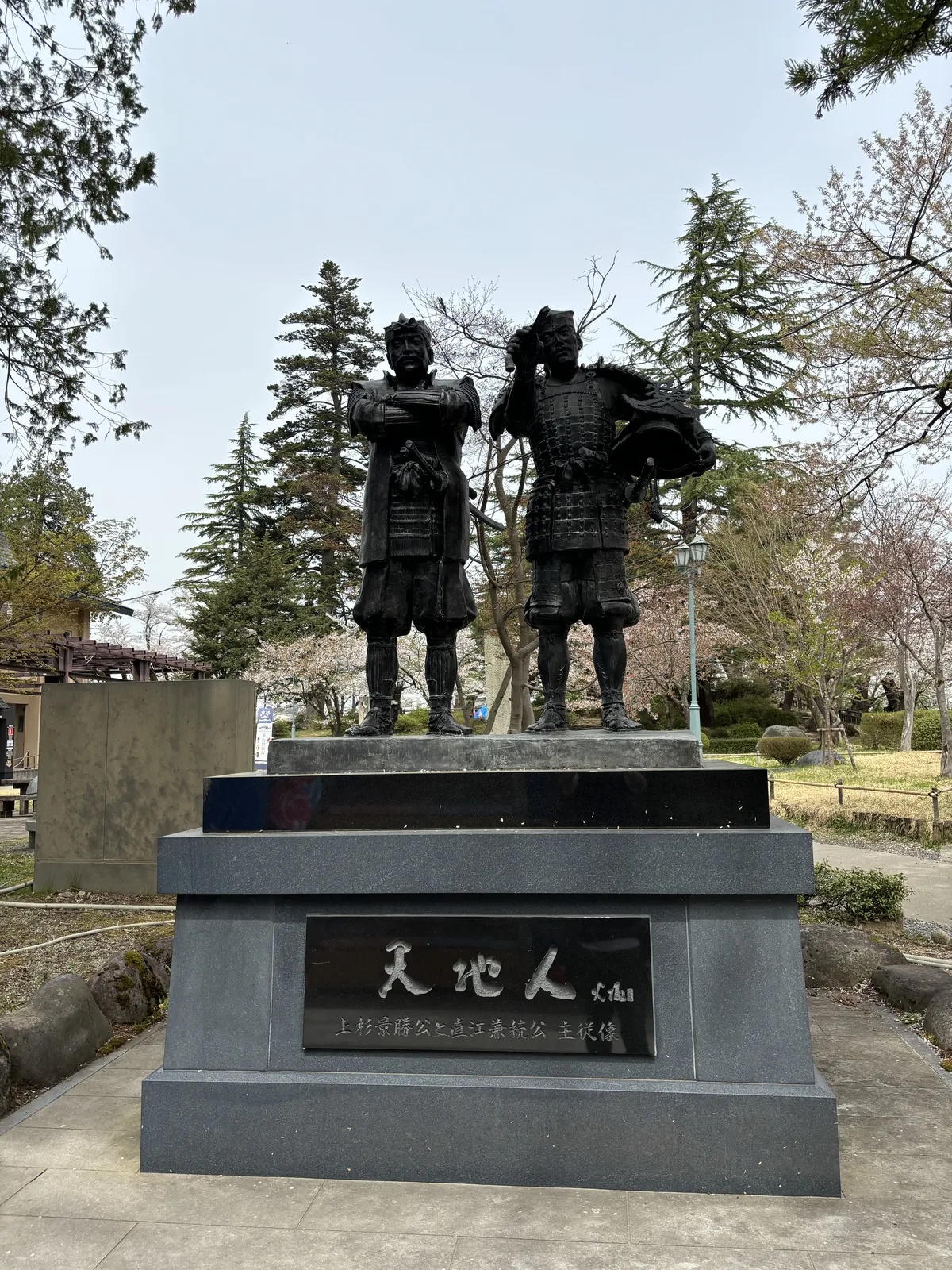 黑御影石的台座上兩尊立像並肩，左邊一位雙手捧著什麼，右邊一位右手扶著頭盔的鍬形。台座正面的銅板刻「天地人」，下方小字「上杉景勝公と直江兼続公　主従像」，即上杉景勝與直江兼續的主從像。背後是杉與正在開的櫻。