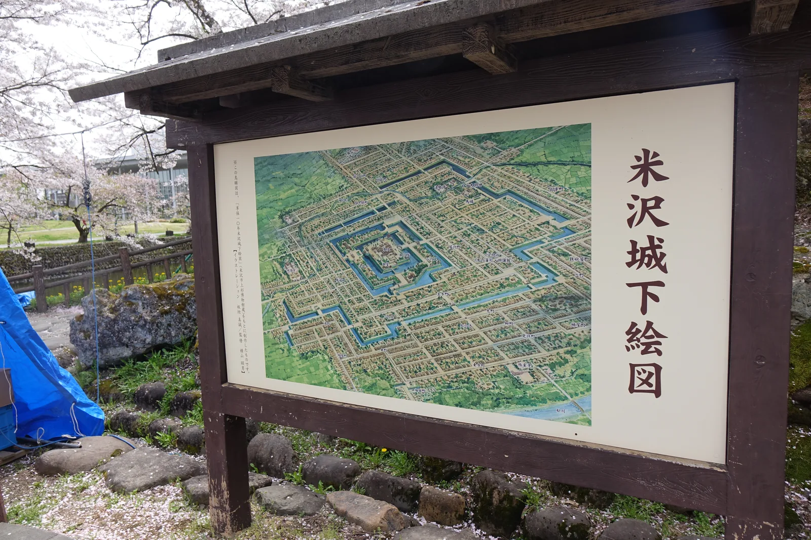 屋根付木框裡一幅彩色的城下町復元圖，圖名「米沢城下絵図」直排在右側。圖上三重方形的水堀一層套一層，中央是本丸，外圈是格子狀的町割。前景是碎石地與剛開的櫻，左側地上蓋著一塊藍色帆布。