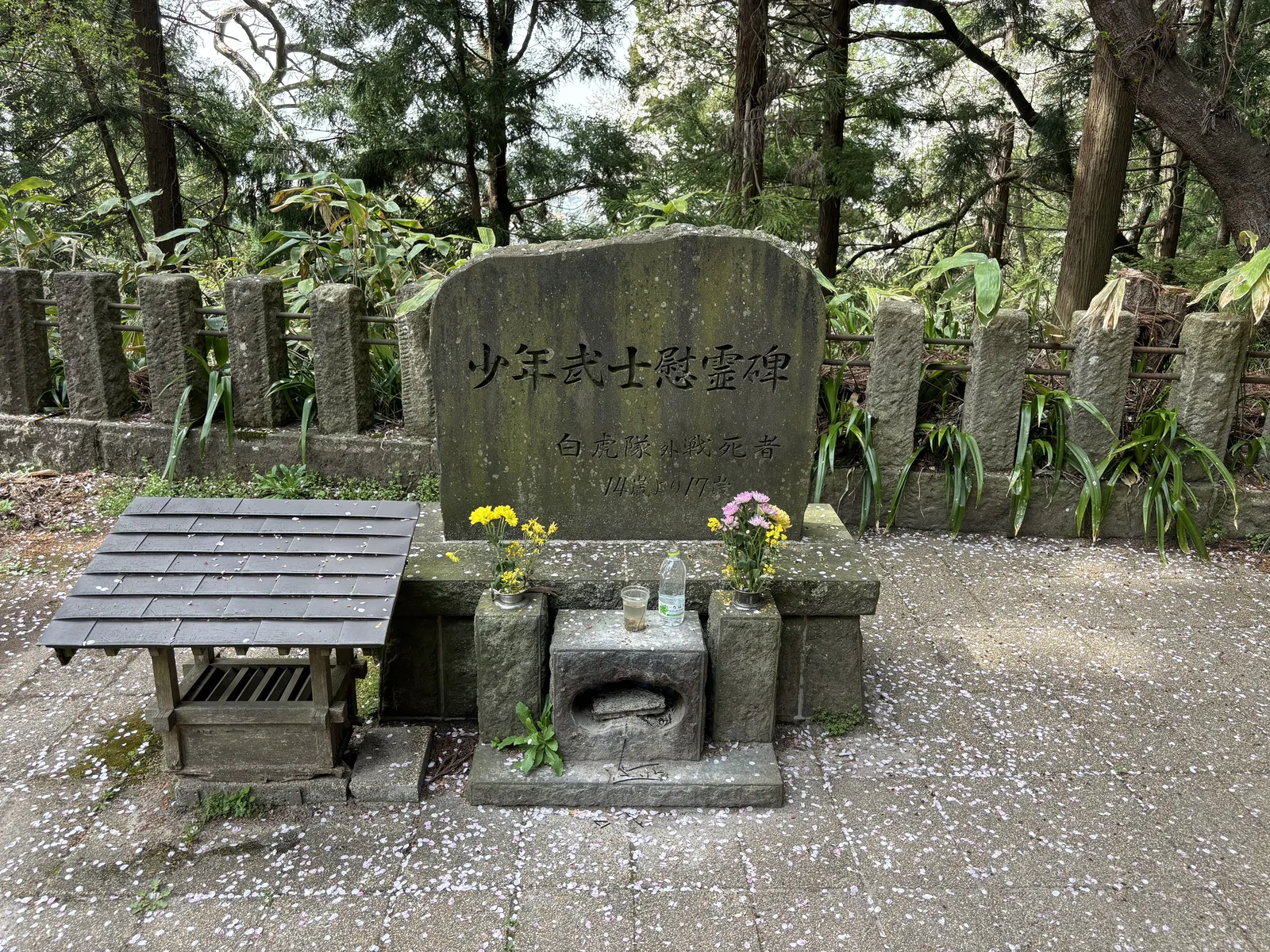 一塊帶弧頂的青灰色石碑，正面橫刻「少年武士慰靈碑」，下方兩行小字。碑前一座石造的供台，插著黃色與白色的菊，左側是一張木格的供物台。碑後是一整排並列的細長墓標與竹叢。