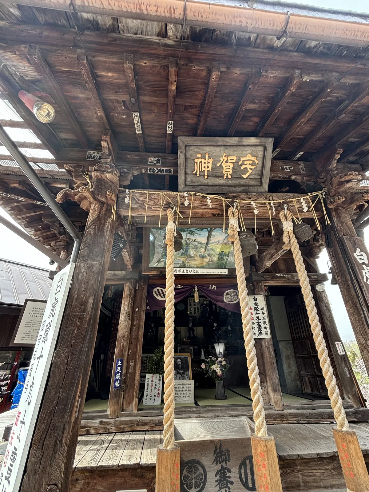 從堂前抬頭看。黑底金字的扁額題「宇賀神堂」，額下橫著一條粗注連繩，垂下兩條同樣粗的鈴緒與白色的紙垂（しで，shide，折成鋸齒狀的白紙條）。堂內昏暗，正面看得到「白虎」開頭的直幟。明治三十七年從三匝堂搬過來的十九士靈像就在裡面。