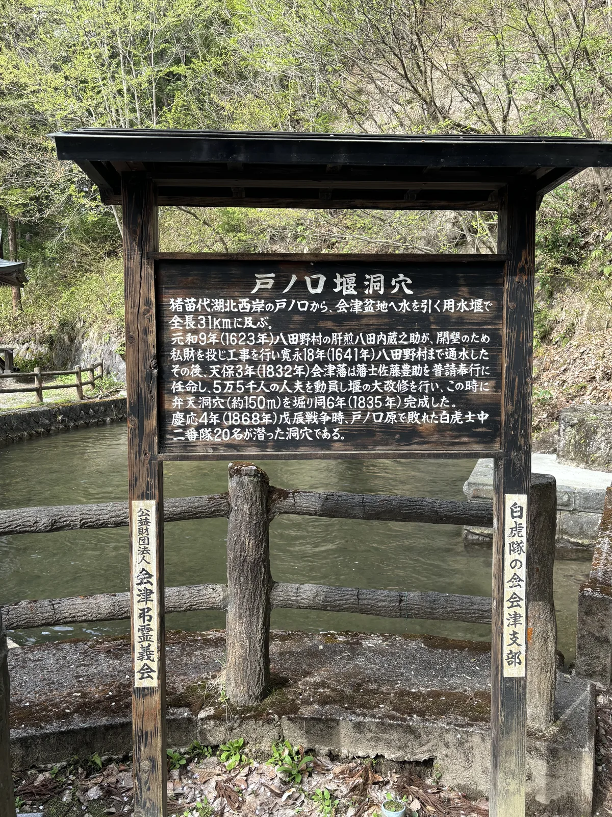 黑色木框與黑屋頂下一面木板，白色字，標題「戸ノ口堰洞穴」。內文記這條用水堰從豬苗代湖北西岸的戶ノ口引水到會津盆地、全長三十一公里，元和九年八田內藏之助自費開工，天保三年會津藩動員五萬五千人大改修，弁天洞穴約一百五十公尺於天保六年完成。