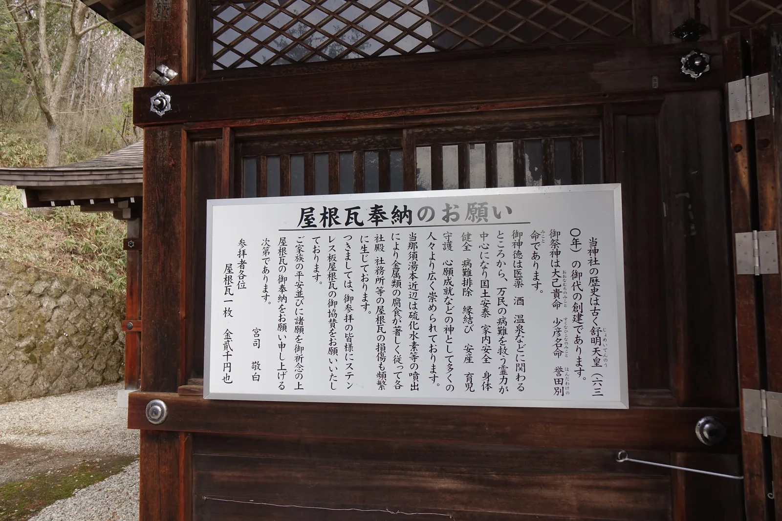 深色木門上掛著白底的告示板，標題「屋根瓦奉納のお願い」。內文自創建年代與祭神寫起，接著說明這一帶因硫化氫噴出使金屬腐蝕嚴重、屋瓦頻繁損壞，請參拜者協贊不鏽鋼板屋瓦；末行寫「屋根瓦一枚　金貳千円也」。