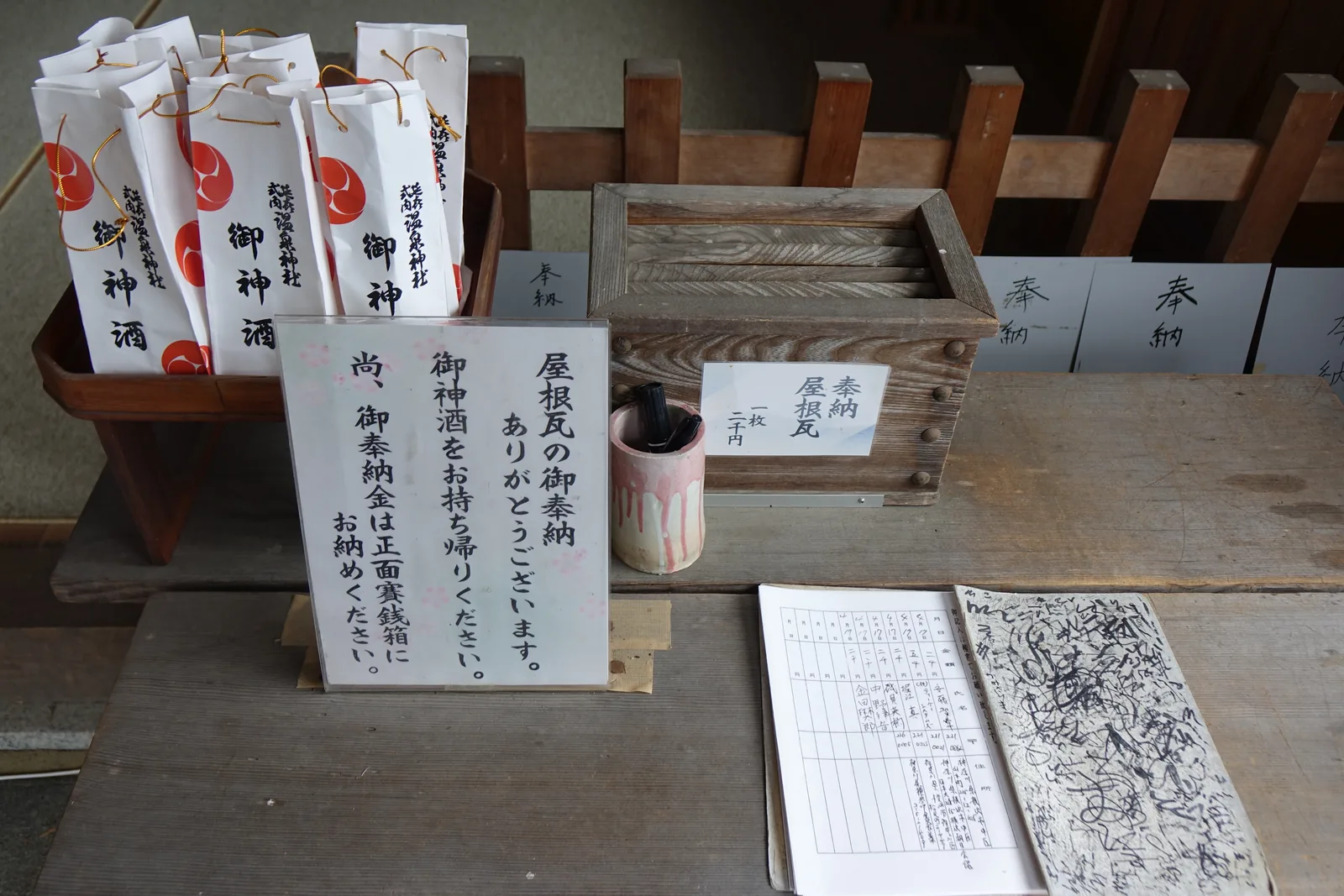木桌上擺著一只舊木箱，貼紙寫「奉納　屋根瓦　一枚二千円」，旁邊是筆筒與翻開的簽名簿，一行一行記著日期、姓名與金額。左側木架上插著一排白紙袋，印紅色三巴紋與「御神酒」。中間的壓克力立牌寫：捐瓦的人請把御神酒帶回去，捐款投進正面的賽錢箱。