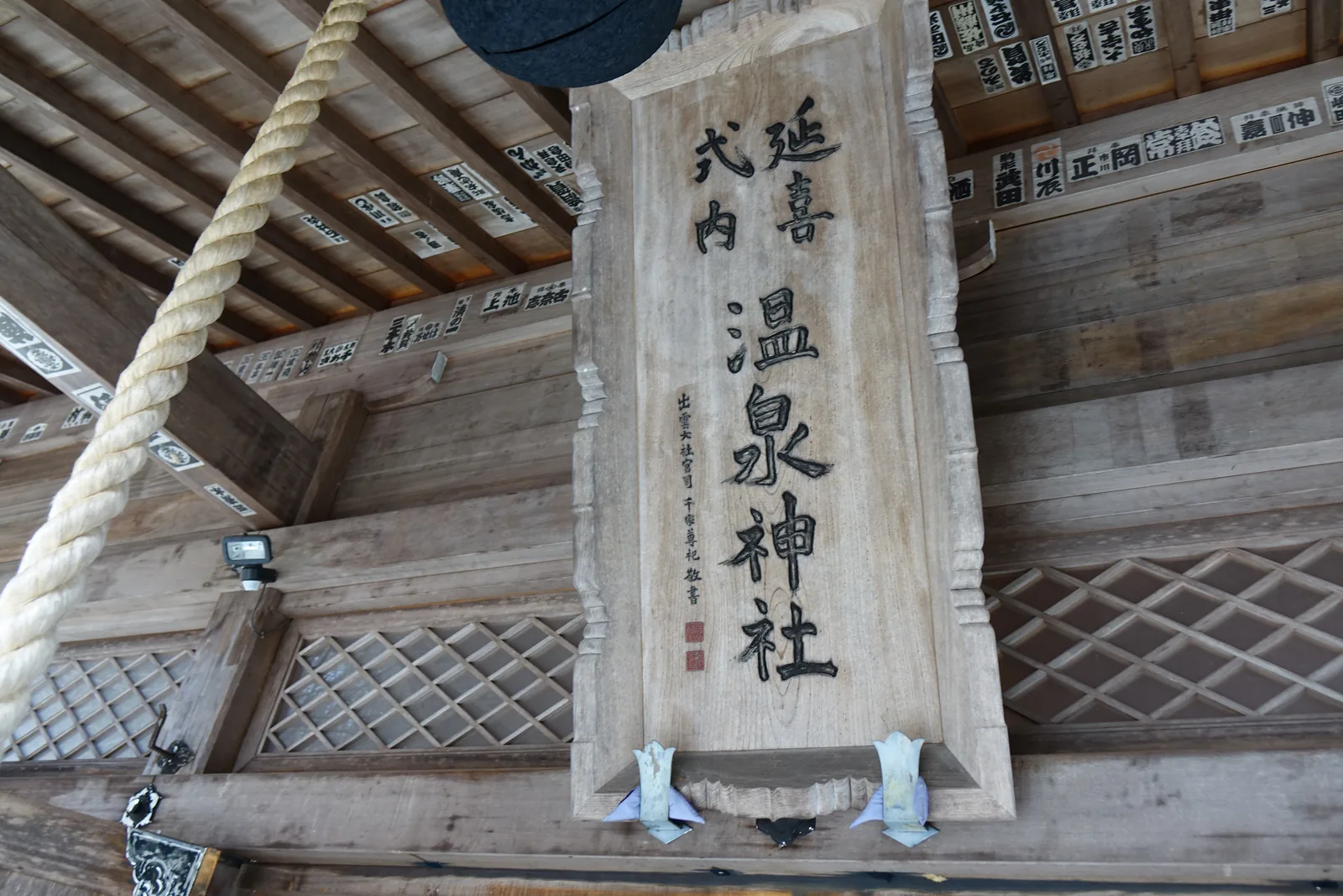 仰角。一面素木扁額掛在拜殿的樑下，刻「延喜式内　温泉神社」，左下小字落款「出雲大社宮司　千家尊祀　敬書」並蓋朱印。扁額周圍的樑上與椽子之間，滿滿排著奉納者姓名的木札。左側是垂下的白色鈴緒。