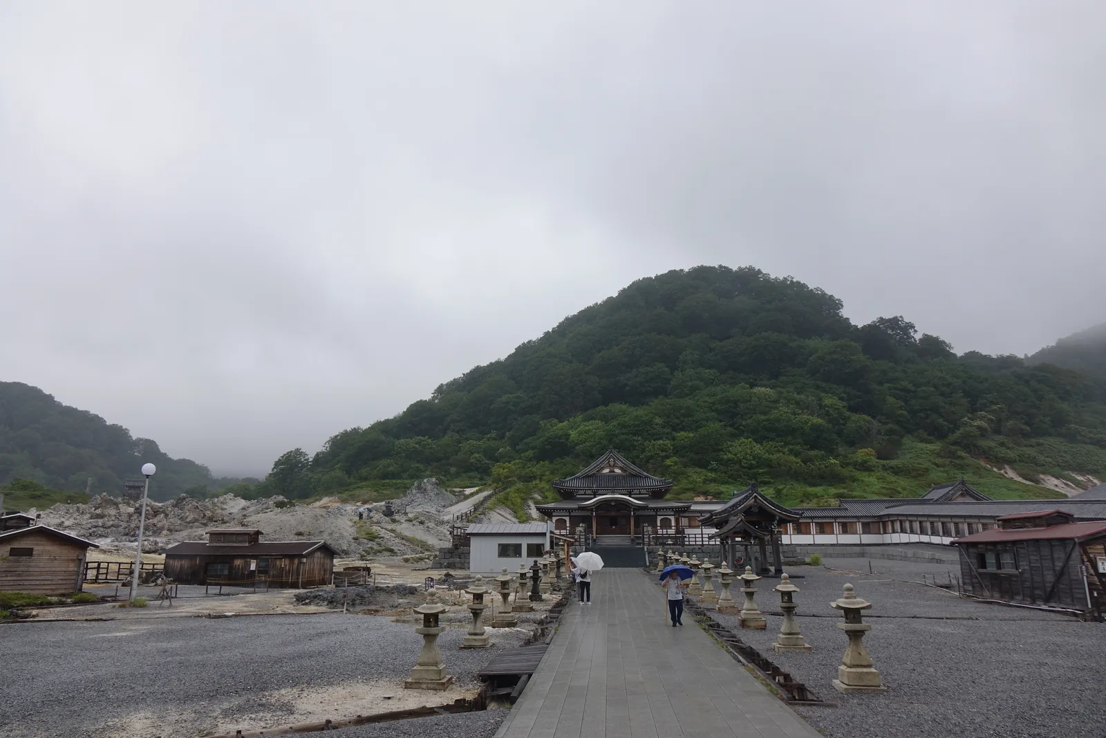 站在山門下往地藏殿看。參道筆直，兩側各一排石燈籠；左邊幾間黑木板的小屋是境內的溫泉，右邊灰白色的碎岩地是被比作地獄的噴氣區。湯、路與地獄擠在同一個畫面裡，這是恐山最準確的一張全景。