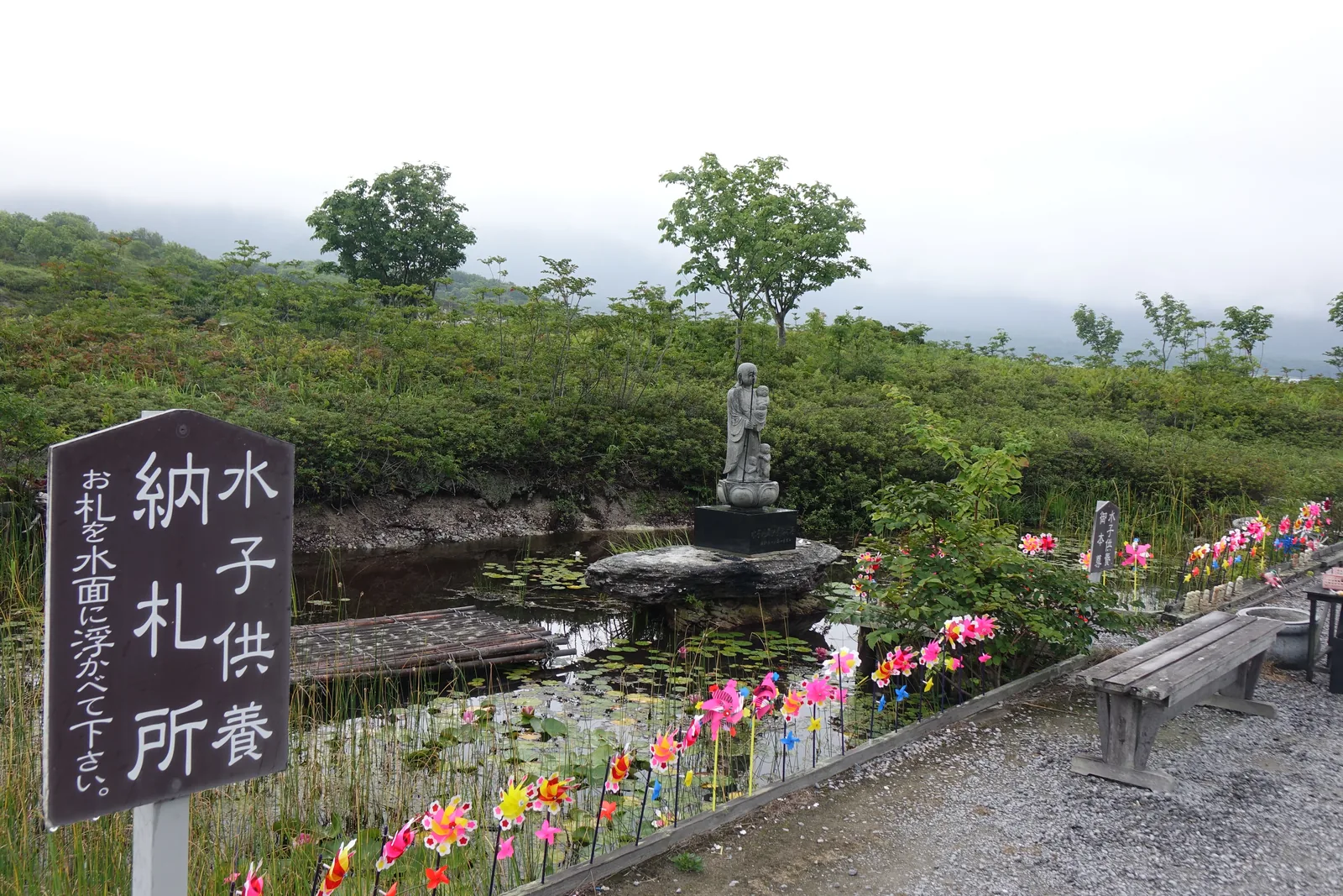 「水子供養納札所」（收受供養水子之納札的地方；納札 nōsatsu 是寫上願文或名字後奉納的紙札）的木牌，小字寫著請把札浮在水面上；牌後是一整排彩色的紙風車，再過去是蓮池與池中一尊立姿地藏。風車本來是給早夭孩子的供物，現在代替鮮花，不限對象。