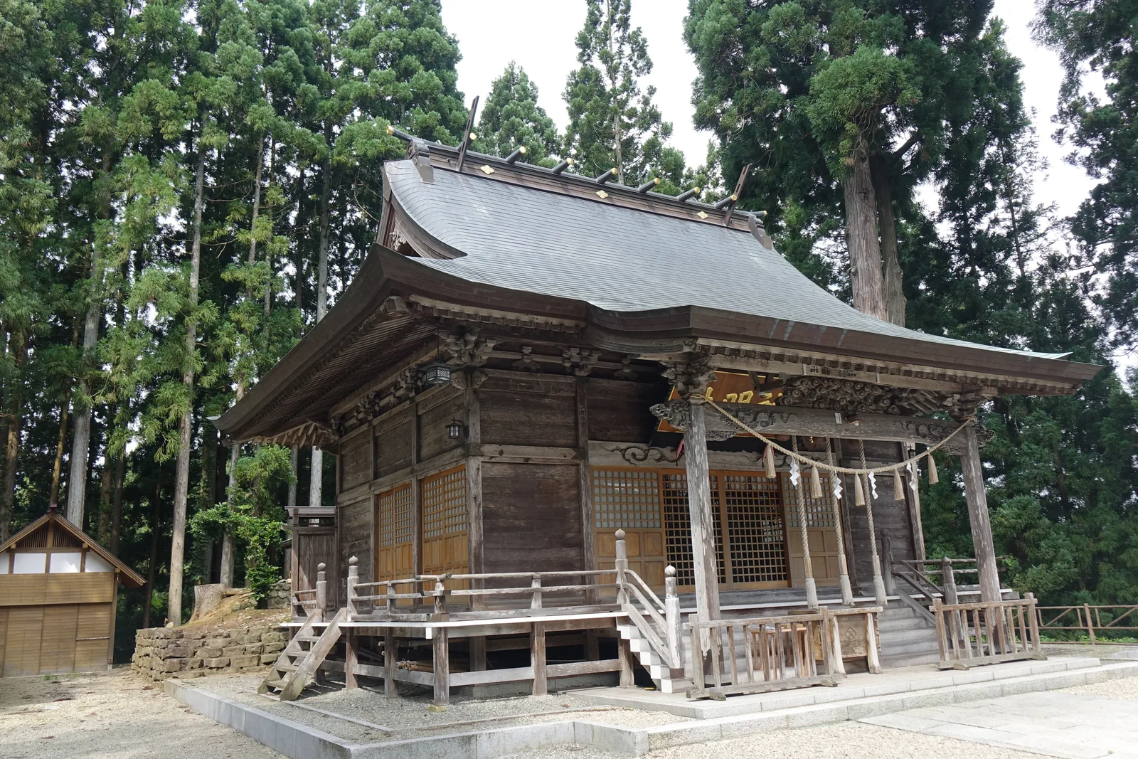 社殿全景。素木（shiraki，不施塗裝的原木）已風化成灰白，入母屋造（irimoya-zukuri，上半懸山、下半廡殿的複合式屋頂）的屋面覆金屬板；建築整體架在高床上，四周繞一圈緣側與高欄（kōran，欄杆），正面出木階。背後全是直上的杉木。