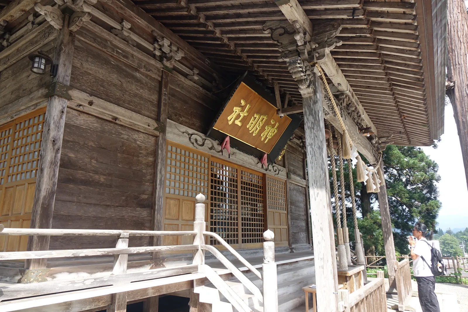 拜殿正面的軒下。黑框金字的扁額題「神明社」，後方是格子戶與白木的高欄，柱與長押全是灰白的素木。鈴緒垂在正中。