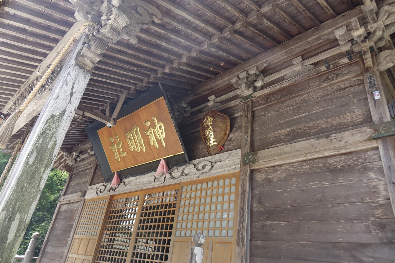 扁額與一只釣燈籠並排。燈籠是金屬製，掛在向拜（gōhai，正面伸出的屋簷）的梁下；再過去是格子戶，門前蹲著一尊小型的狛犬（komainu，成對守在社前的獅形石像）。