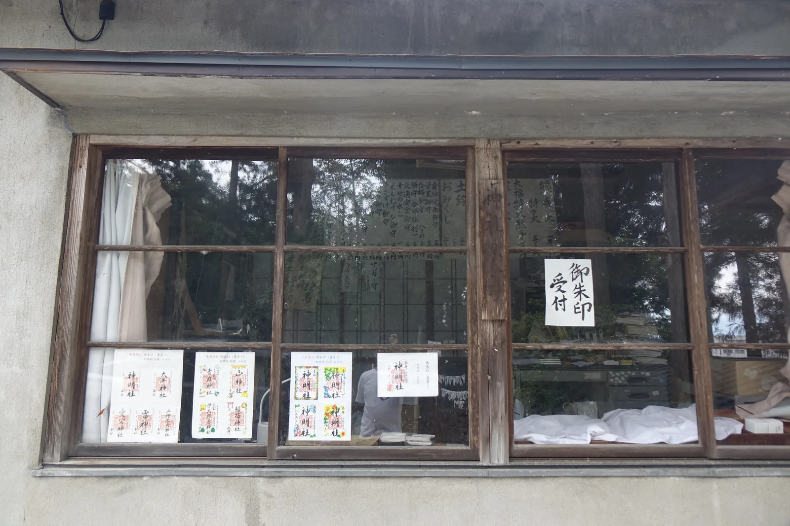 社務所的木框玻璃窗，貼著手寫的「御朱印受付」。窗台上排著幾種御札（gosatsu，神社授與的護符）與御守，室內掛著寫滿名字的祈願木牌。