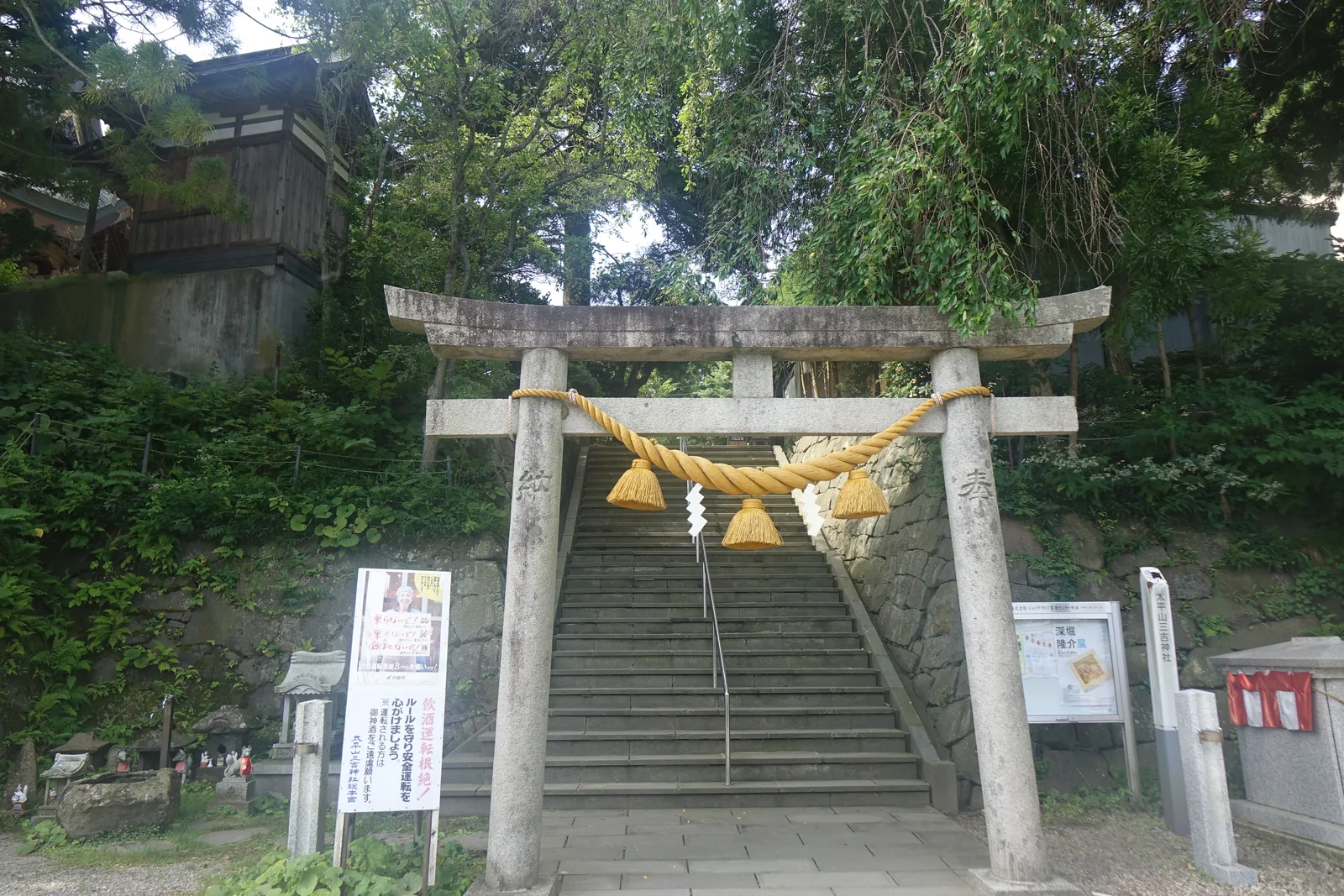 正參道入口的石造明神鳥居（myōjin torii，笠木兩端上翹、帶島木的鳥居形式）。黃色的注連繩垂著四條紙垂（shide，摺成鋸齒狀的白紙）；鳥居後面是石階，兩側石垣夾道。左右立著掲示板與社號標（shagōhyō，刻著社名的石柱）。