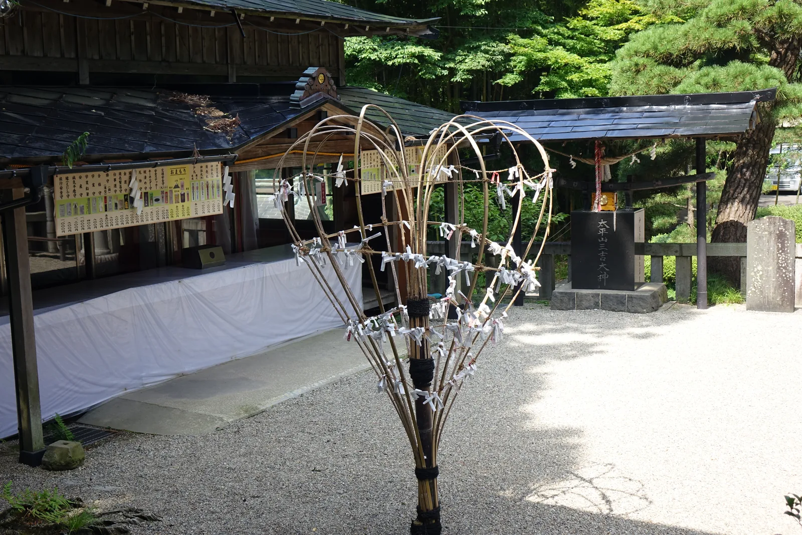 砂礫地上立著一具只剩骨架的祭具：中心一根棒，竹篾自頂端彎成弧形向四方張開，篾上繫滿紙垂。神社對梵天的說明正是「用色彩豐富的布與錦裝飾竹籠」——去掉布，剩下的就是這個形狀。右後方是刻著「太平山三吉大神」的黑色石碑。