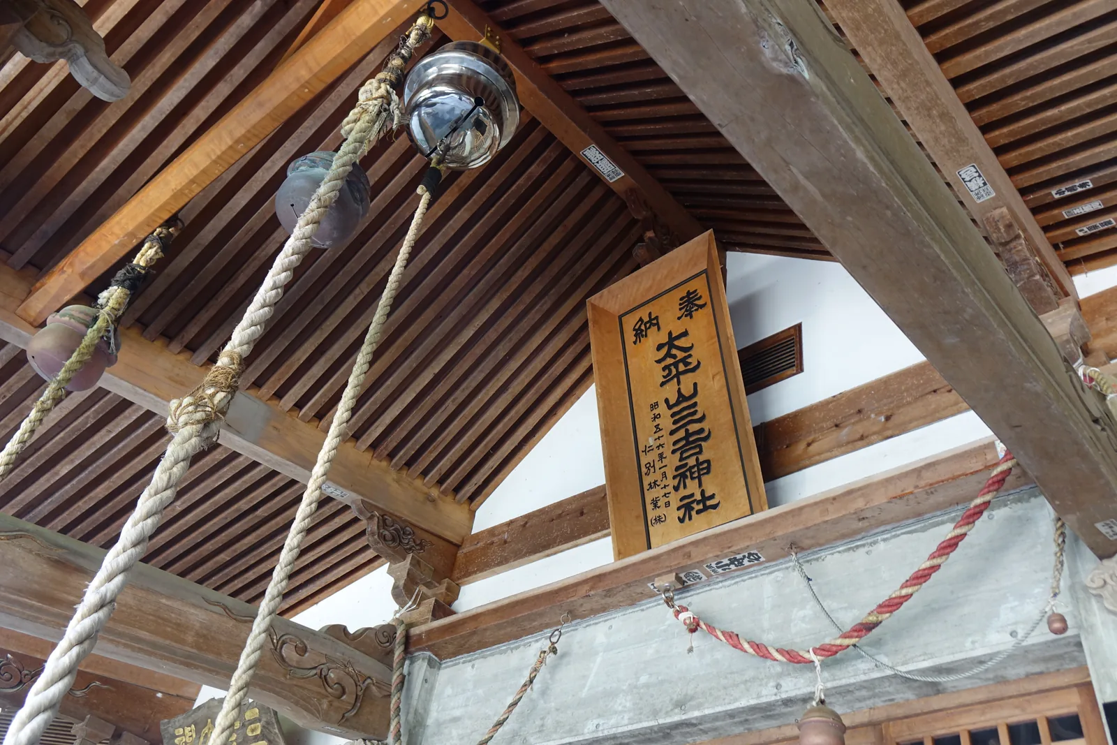 拜殿內部仰視。化妝屋根裏（keshō-yaneura，不做天花板、直接讓椽木外露）的垂木（taruki，屋頂下方的椽）排成一片，幾束鈴緒垂下來；正中立著一塊木札，寫「奉納　太平山三吉神社　昭和五十六年一月十七日　仁別林業株式會社」。一月十七日就是梵天祭。